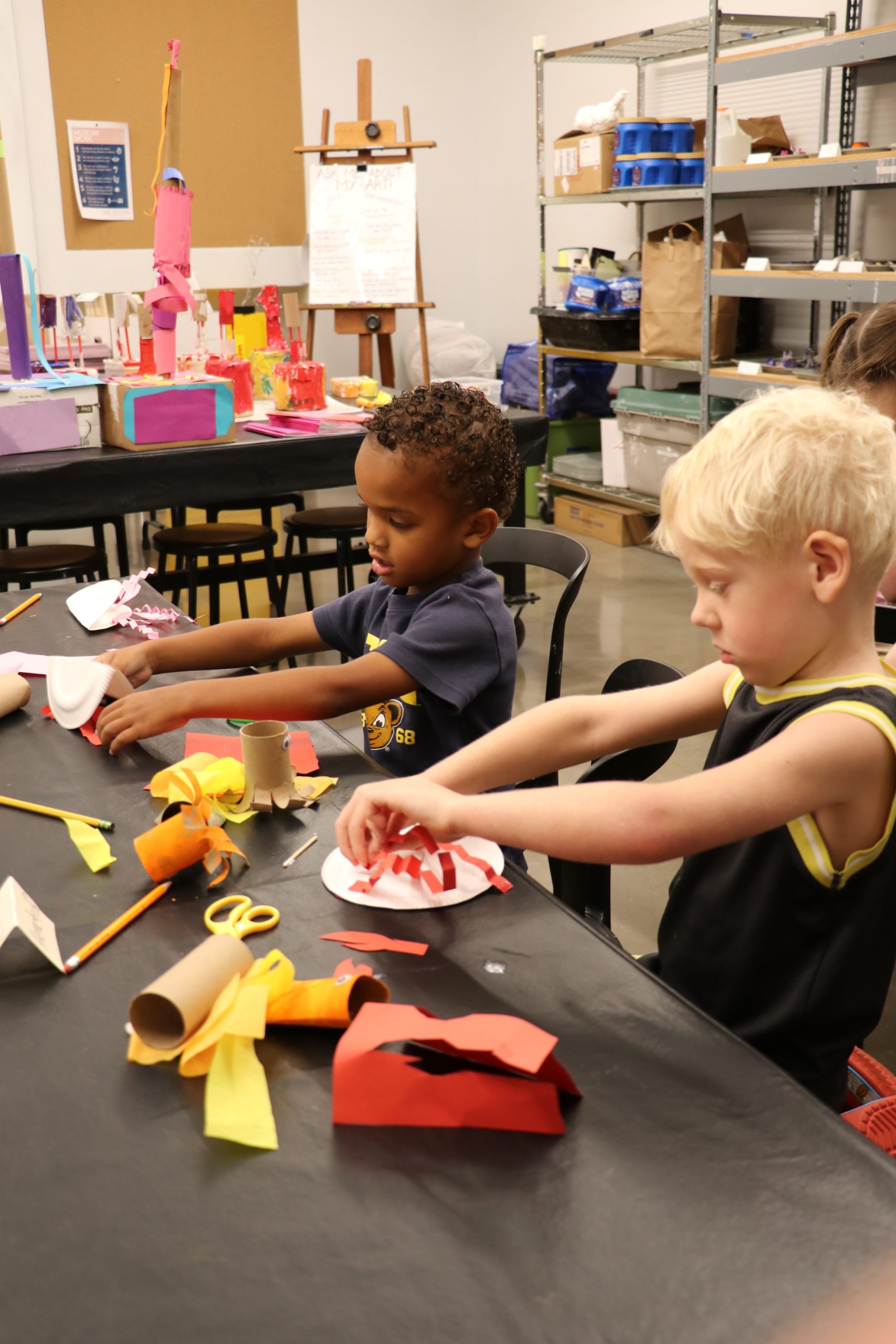 Two young boys sit at a table doing arts and crafts with colored paper, pencils, and other materials in a classroom with shelves and art supplies in the background.