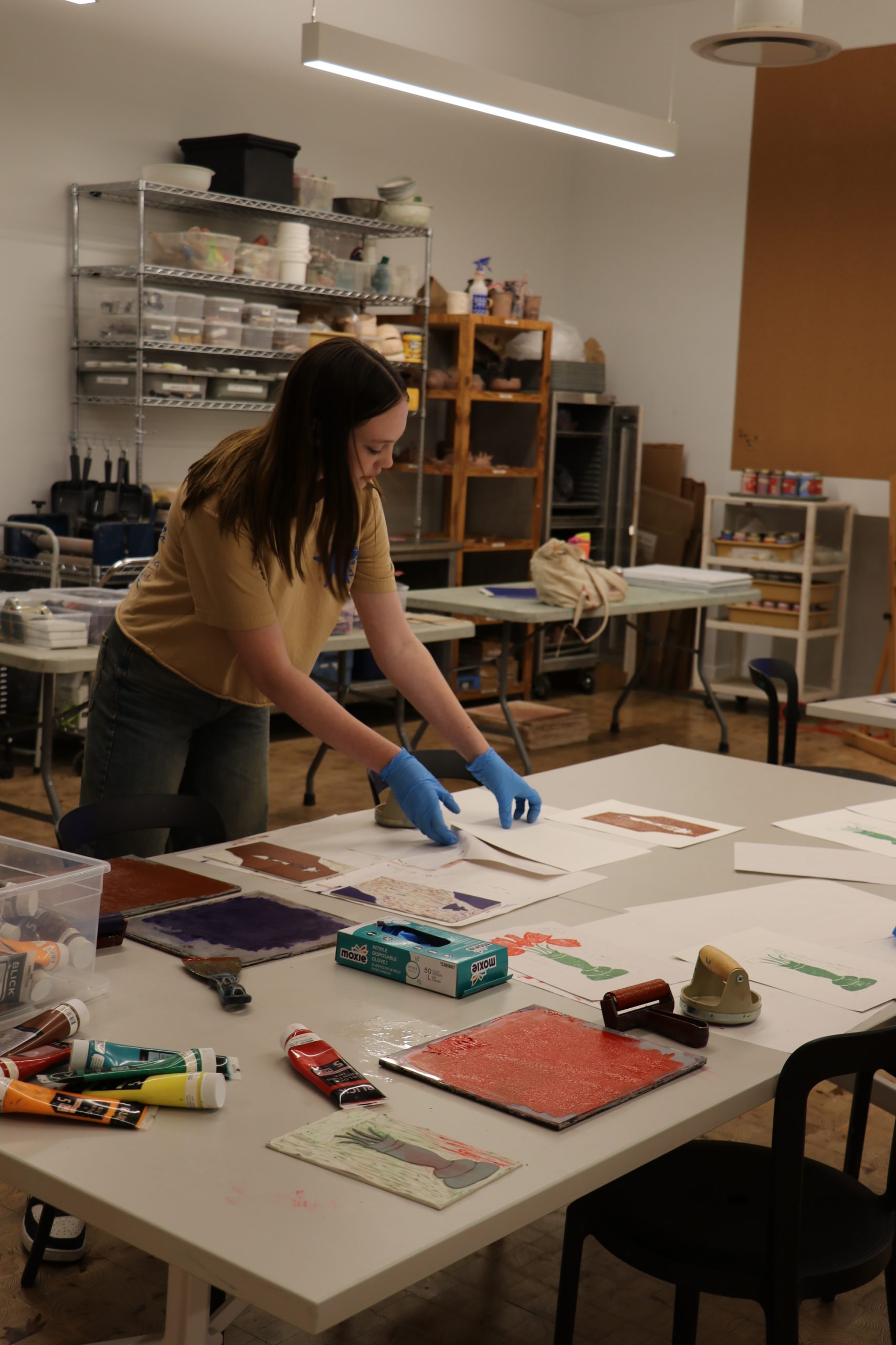 A person wearing blue gloves arranges papers on a table covered with art supplies in a workshop or studio with shelves and materials in the background.