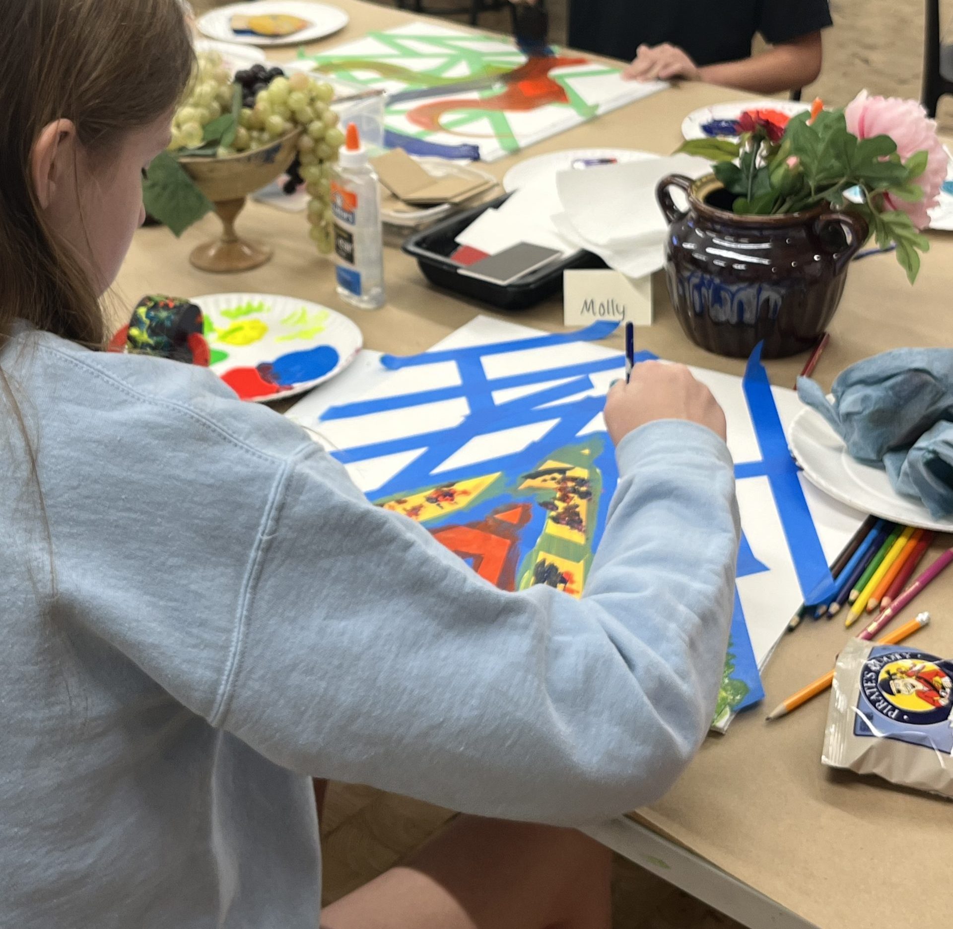 Two girls sit at a table painting abstract art on paper in a classroom. Art supplies, snacks, and a vase with flowers are on the table. One wears a "MEOW" shirt, and the other works on a blue and white design.