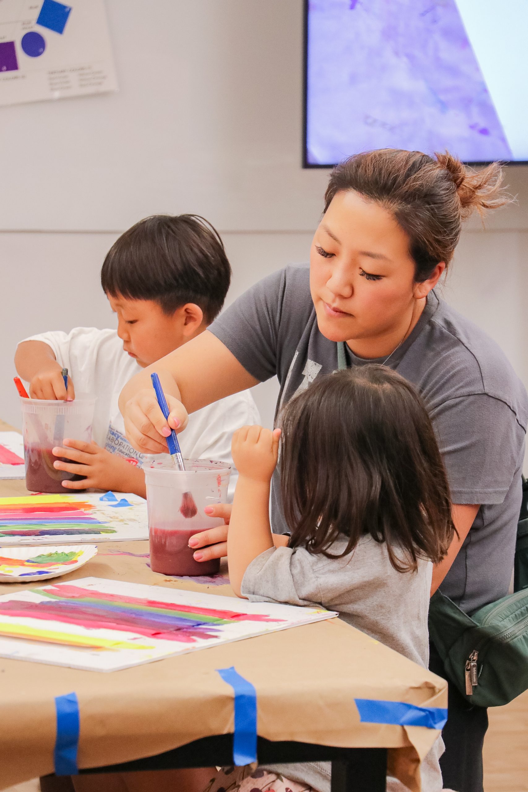 A woman helps two young children paint at a table covered with paper, paintbrushes, and colorful artwork. The children are focused on their painting, and the atmosphere appears creative and engaging.