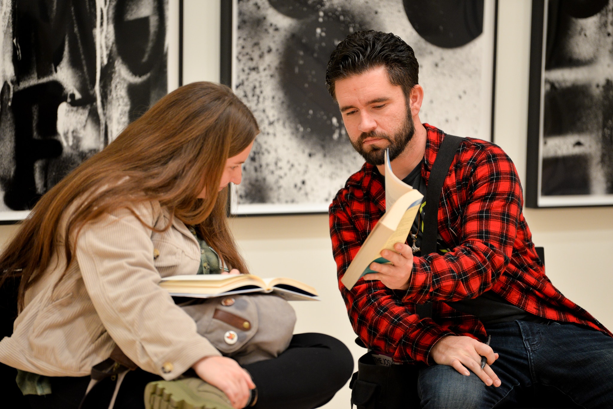 A man in a red plaid shirt shows a book to a woman with long brown hair as they sit together, reading and discussing in front of abstract black-and-white artwork.