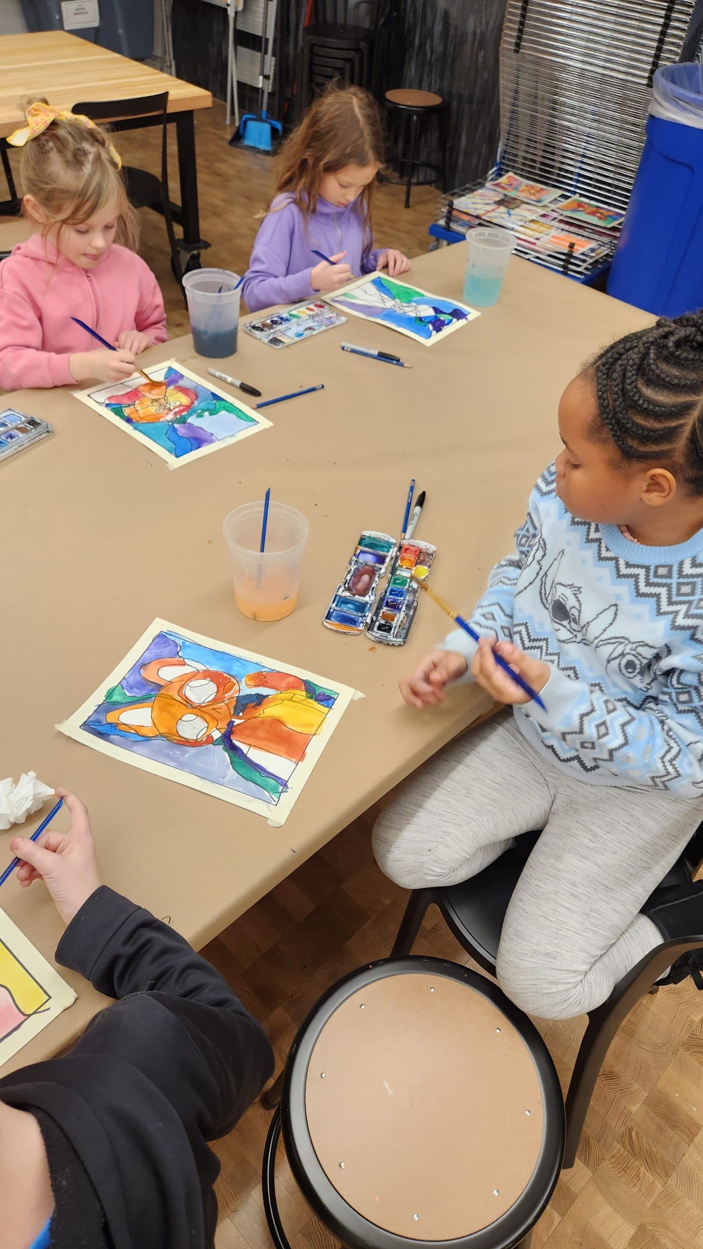 Four children sit around a table, painting colorful pictures with watercolors. Art supplies and cups of water are on the table, and the children are focused on their artwork.