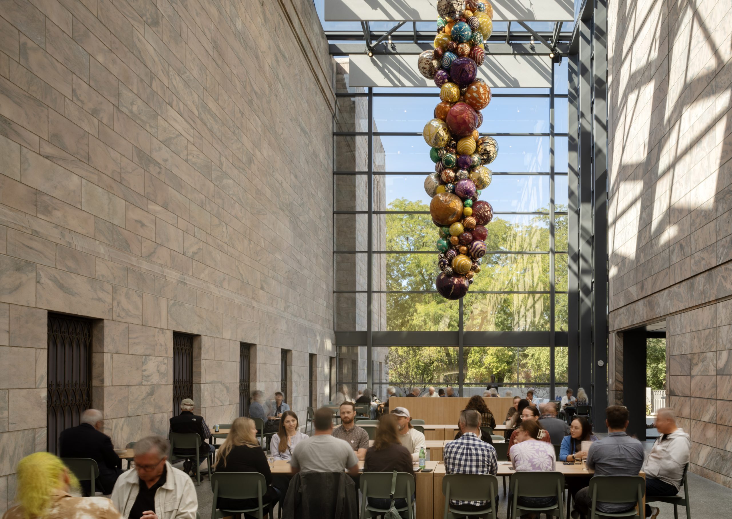 A spacious, modern cafe with high ceilings and large windows letting in natural light. People sit at tables eating and talking. A tall, colorful glass sculpture hangs from the ceiling. Trees are visible outside through the windows.