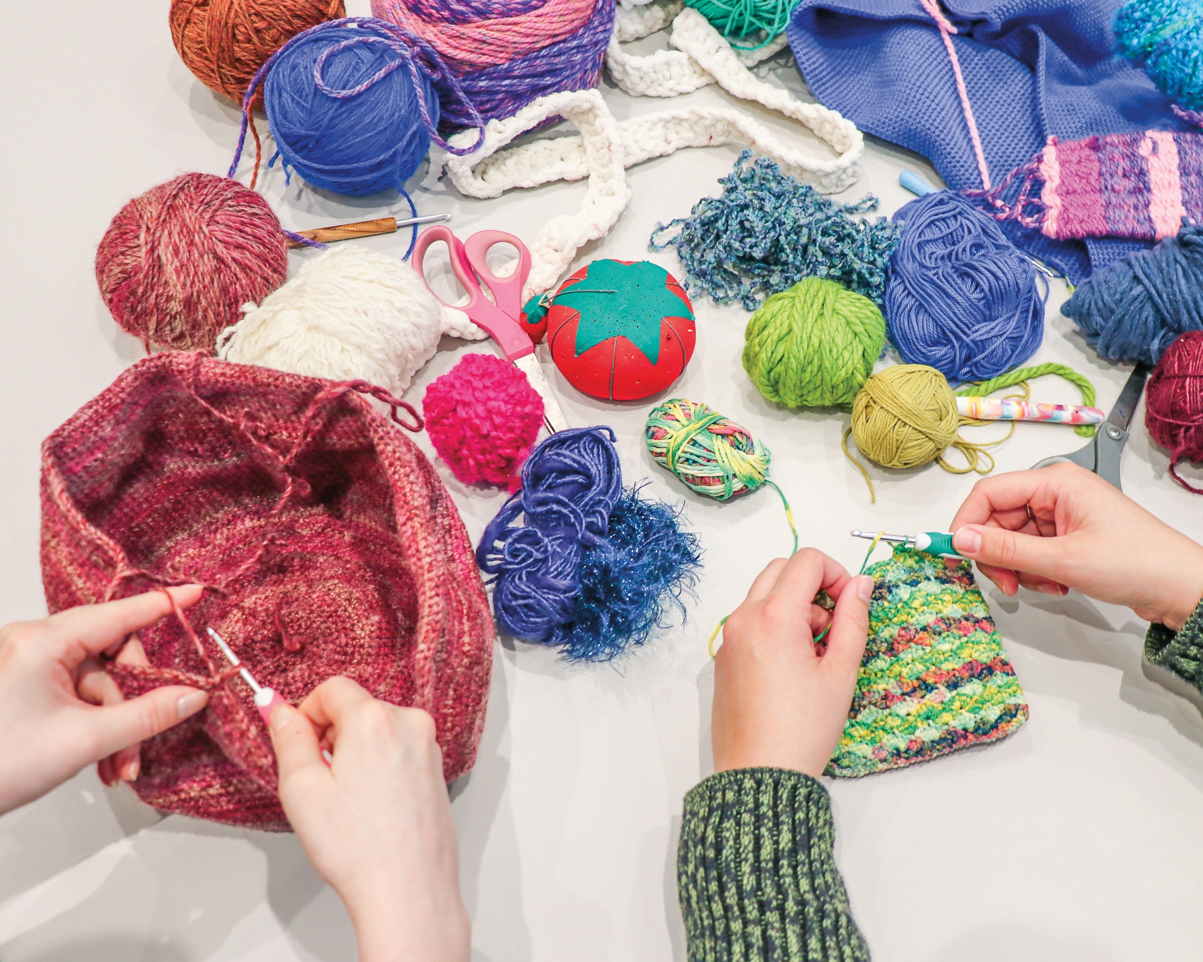 Two people crochet with colorful yarn. Various yarn balls, crocheted pieces, scissors, and a strawberry-shaped pincushion are scattered on a table. One person works on a pink project, another on a multicolored piece.
