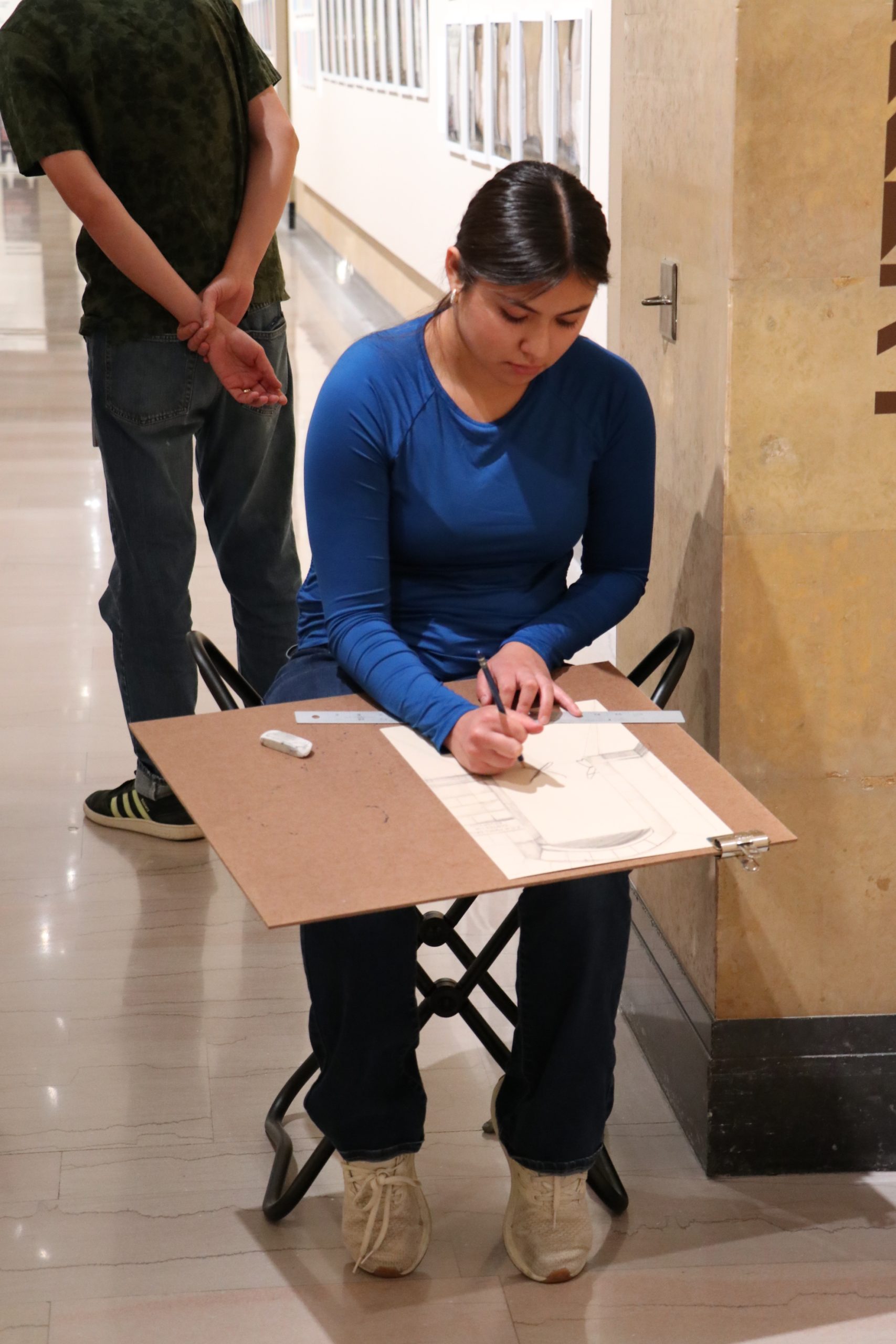 A woman in a blue long-sleeve shirt sits on a small stool, sketching on a drawing board in a hallway. Another person stands in the background with their hands clasped behind their back.