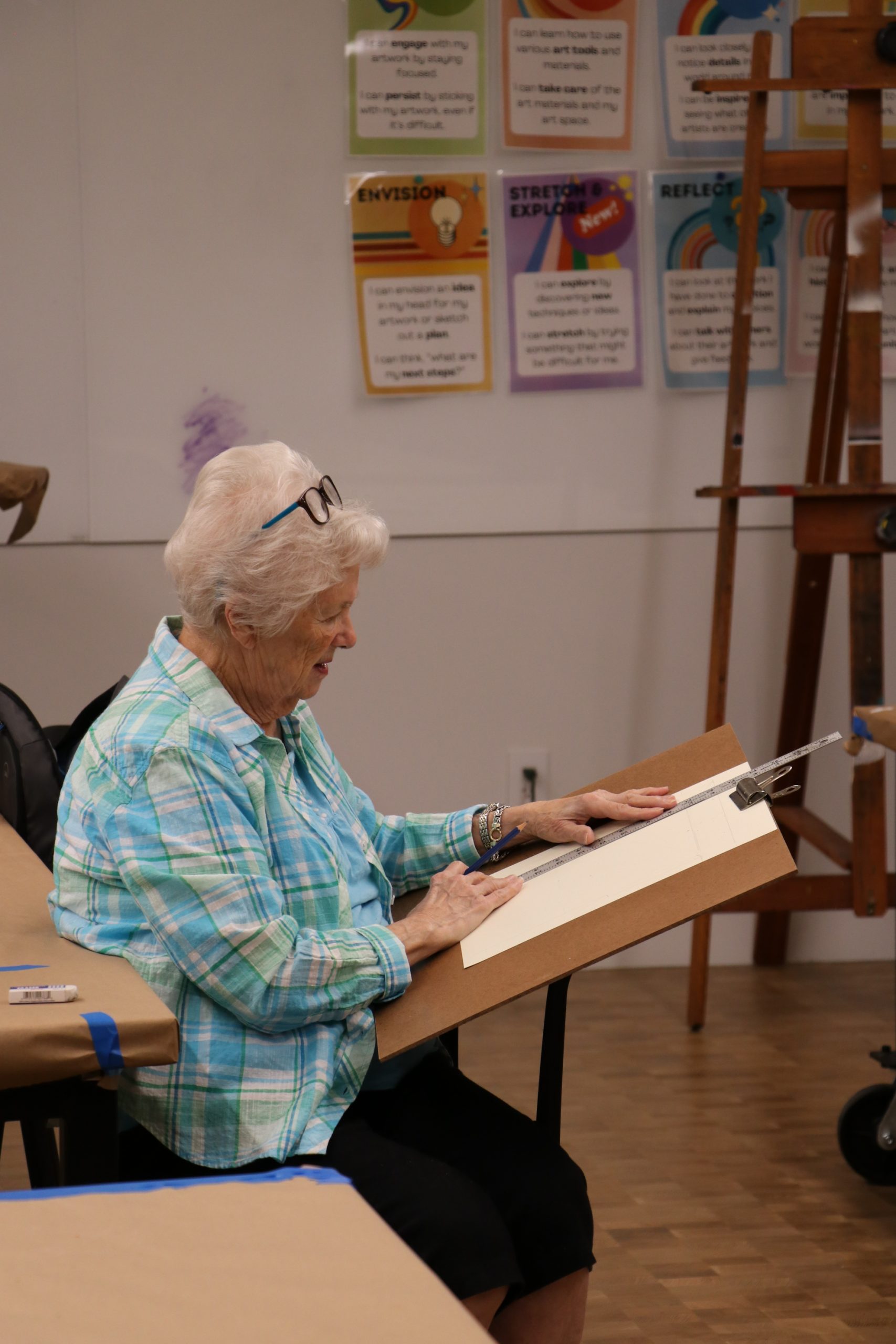 An elderly woman with white hair and glasses sits on a chair, sketching on a drawing board in an art classroom. Colorful motivational posters are displayed on the wall behind her.