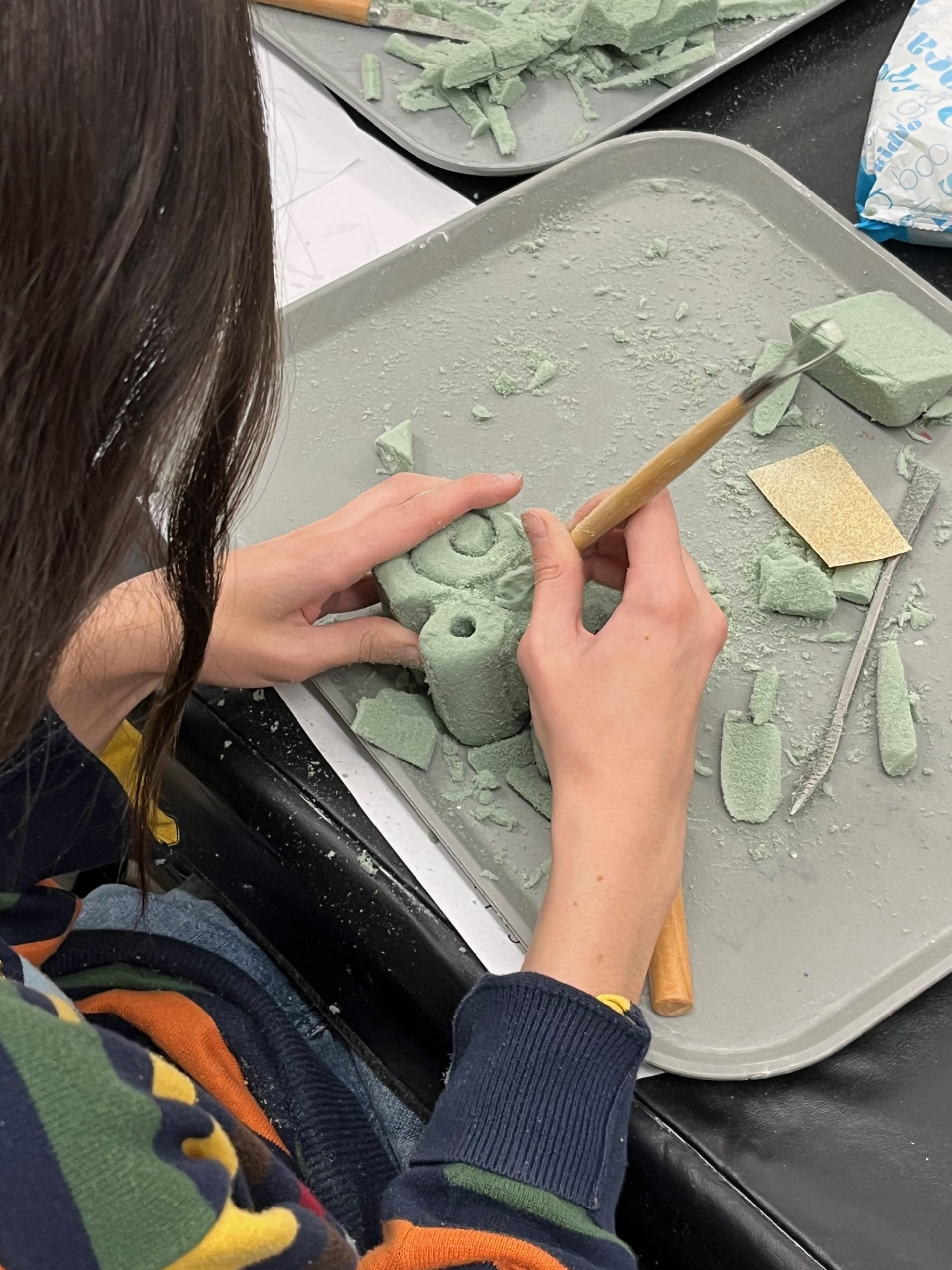 A person carves a small green foam sculpture with a wooden tool while holding it over a tray covered with foam scraps, sandpaper, and other carving tools.