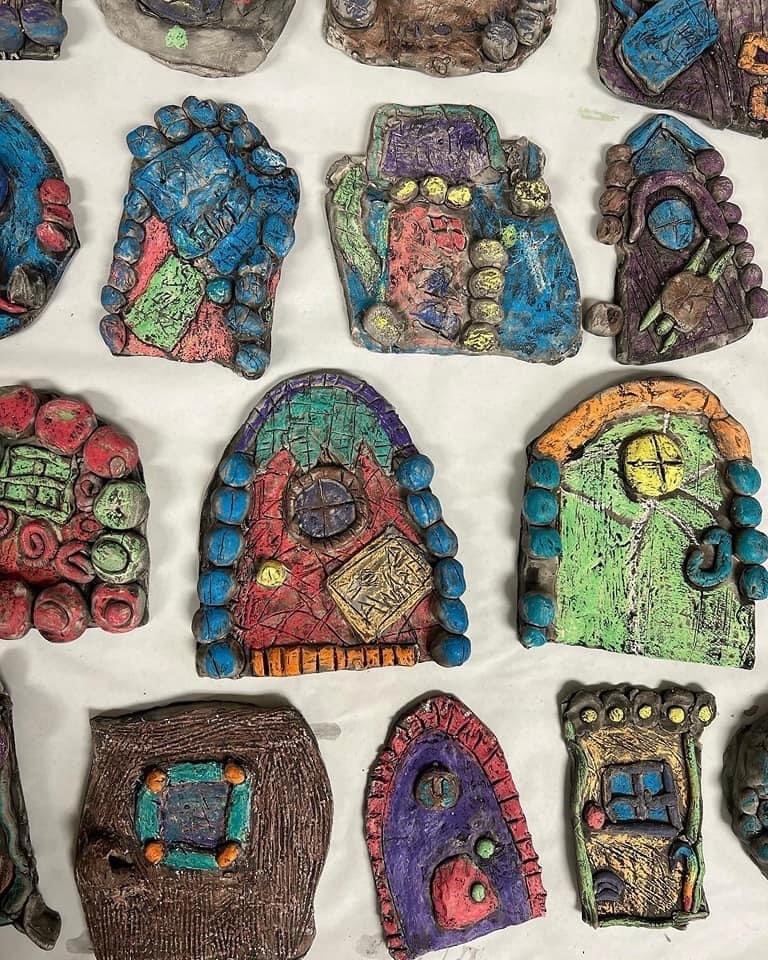 Colorful, handmade clay doors with various patterns, beads, and shapes are arranged on a white surface, each uniquely decorated with vibrant paint and textured designs.