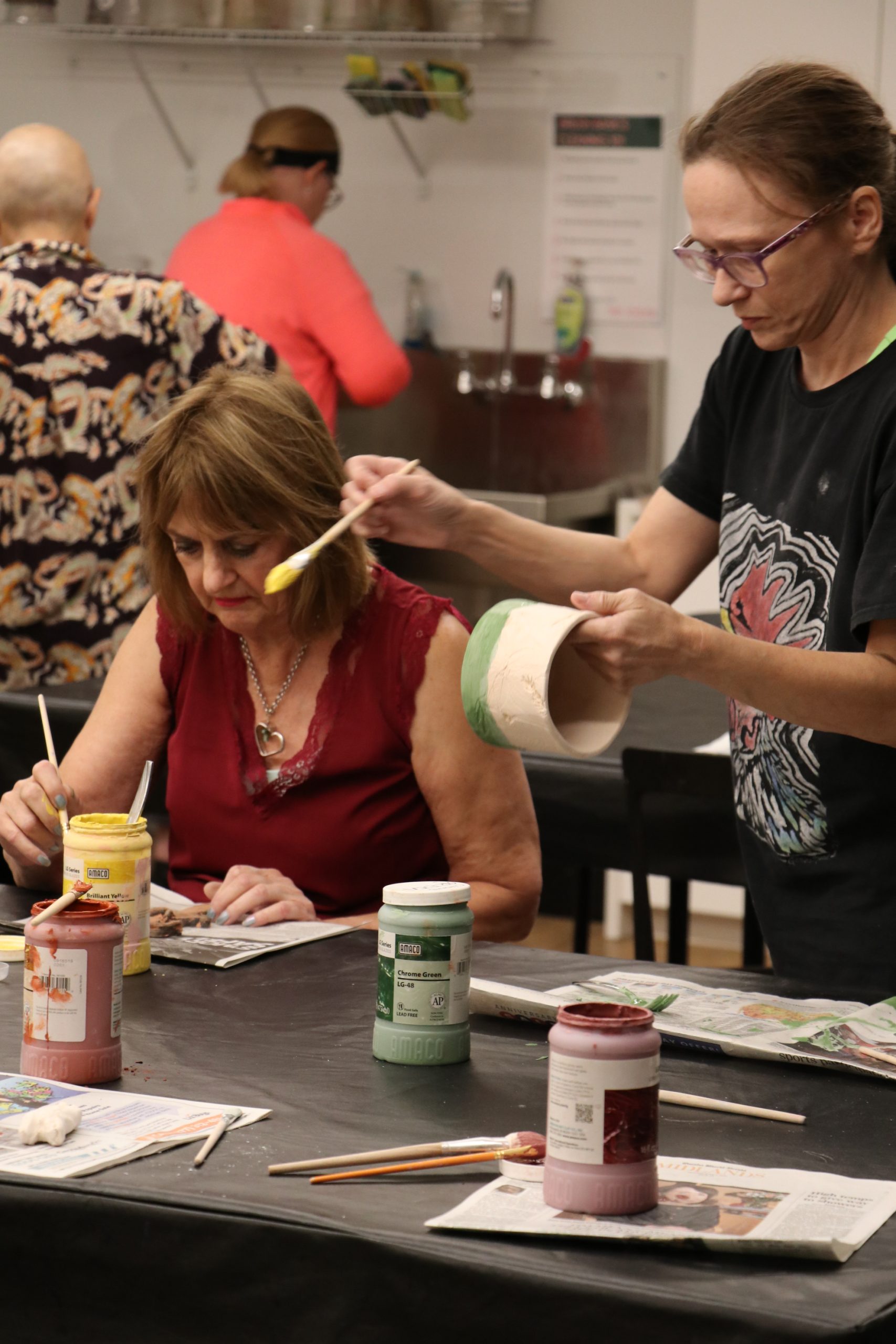 Two women sitting at a table paint pottery with brushes. Various paint jars, brushes, and newspapers cover the table. Other people are visible working in the background of the art studio.