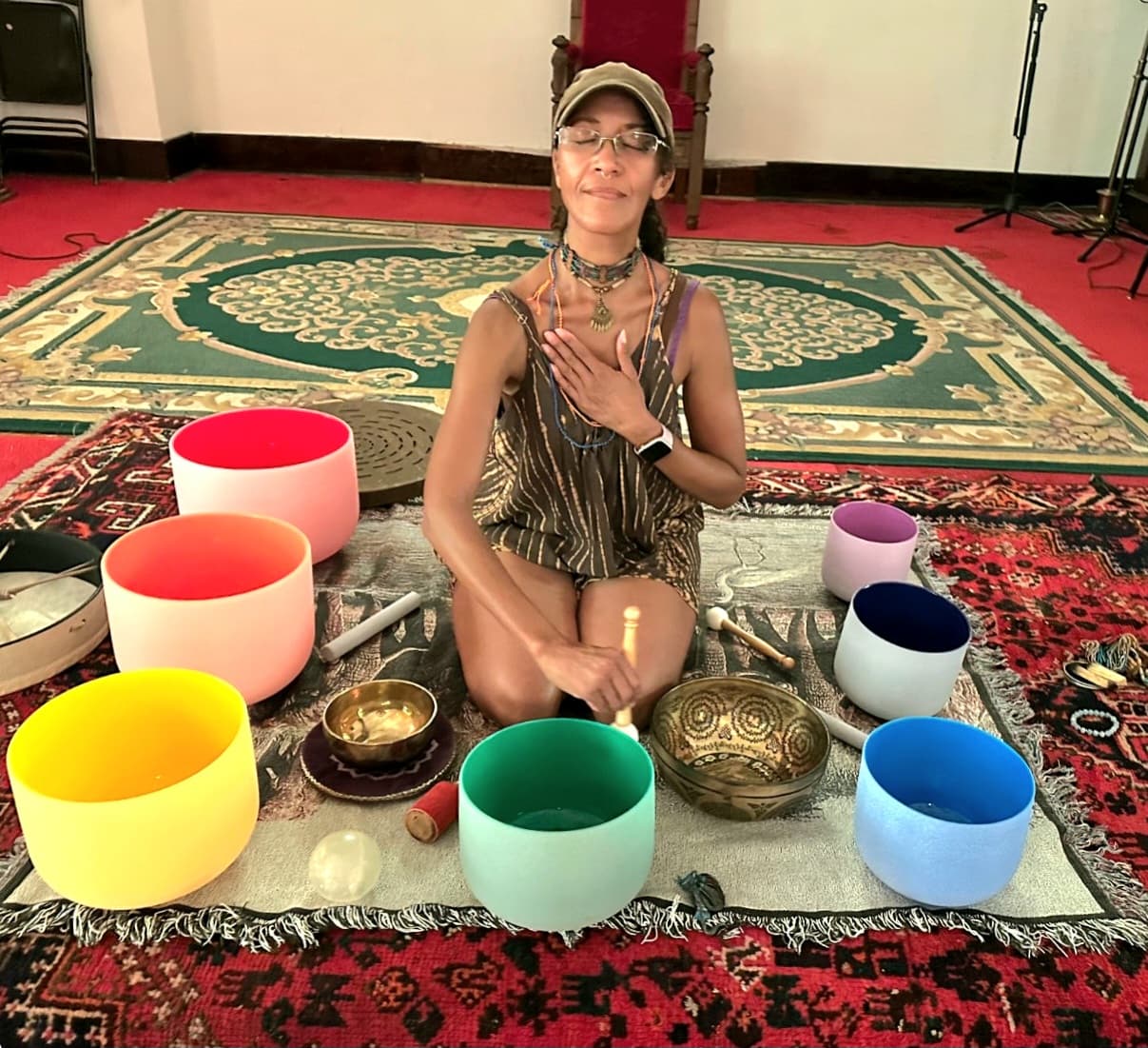 A woman sits cross-legged on a rug surrounded by colorful singing bowls. She holds a mallet in one hand, with her other hand resting on her chest, eyes closed in a peaceful expression. The setting is indoors with ornate carpets.