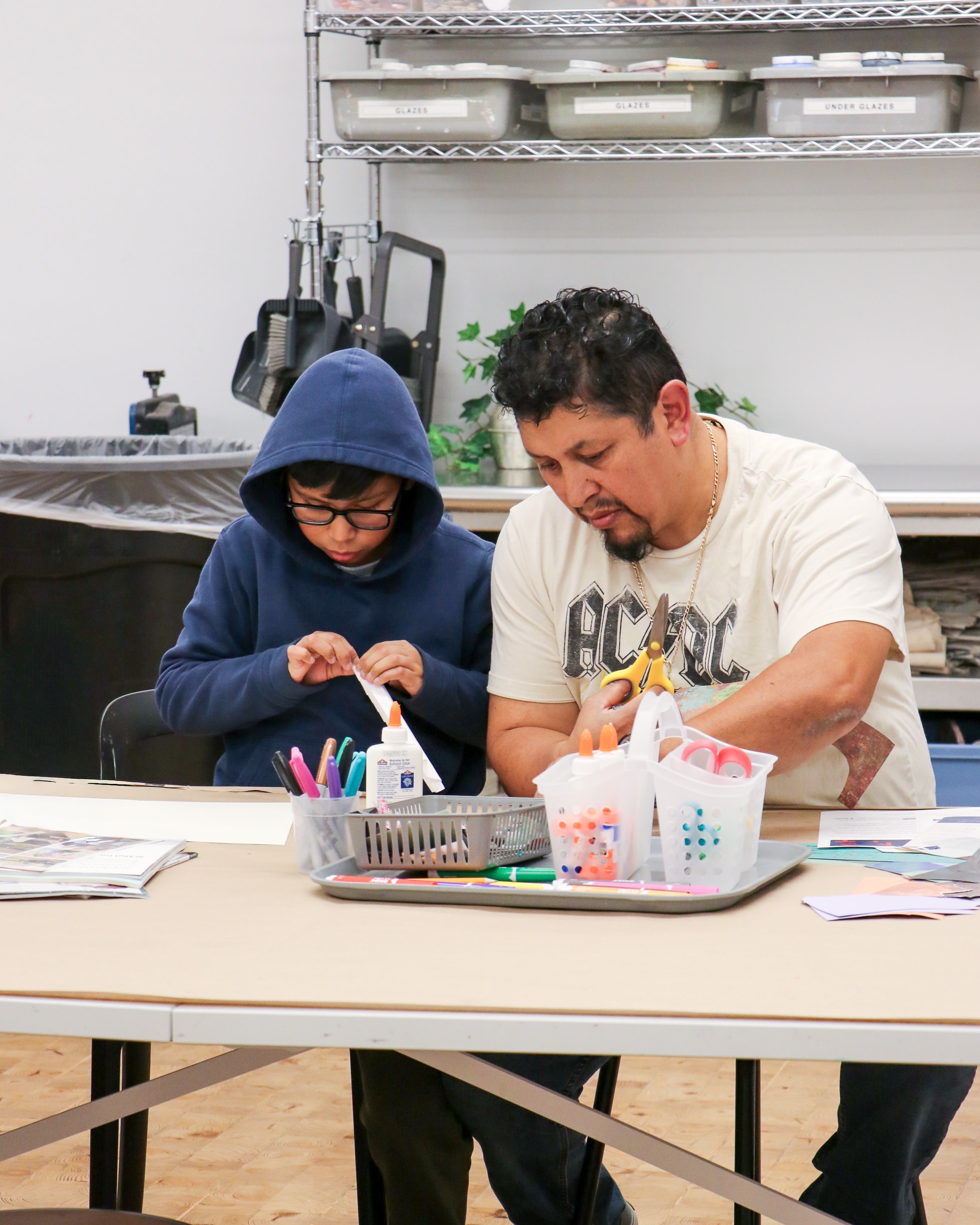 An adult and a child sit at a table doing arts and crafts. The child wears glasses and a hoodie, using glue, while the adult assists. Art supplies, markers, and containers are on the table in front of them.