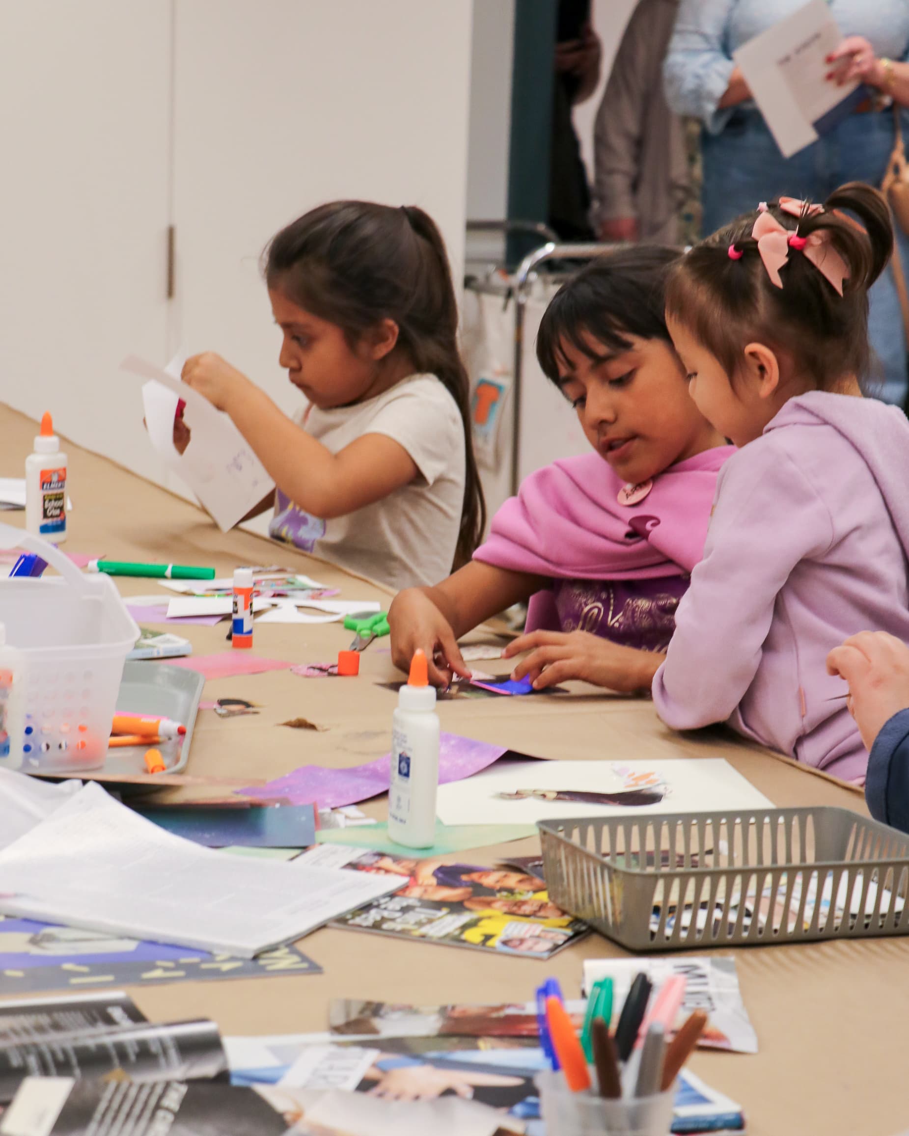 Three young children sit at a table covered with art supplies, including glue sticks, markers, and papers, focused on making crafts together in a classroom or art studio setting.