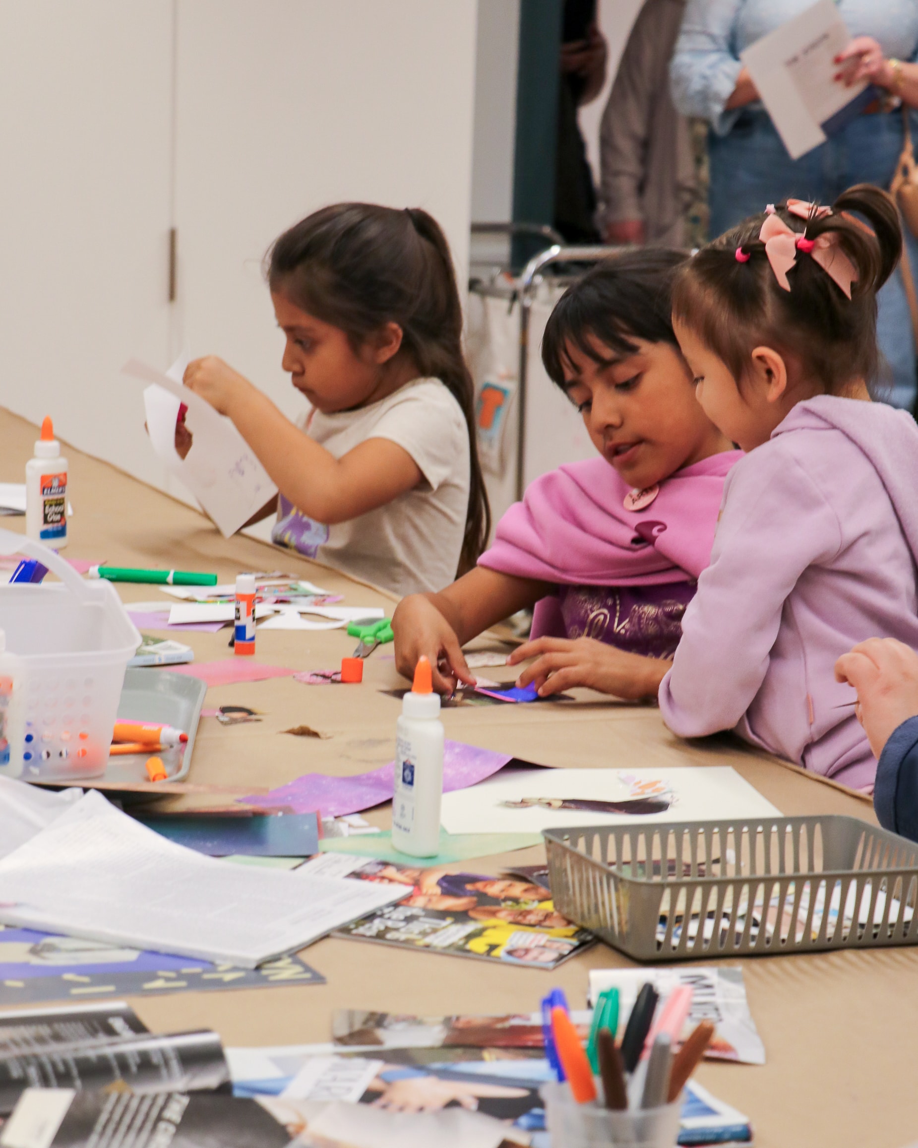 Three young children sit at a table covered with art supplies, including glue sticks, markers, and papers, focused on making crafts together in a classroom or art studio setting.