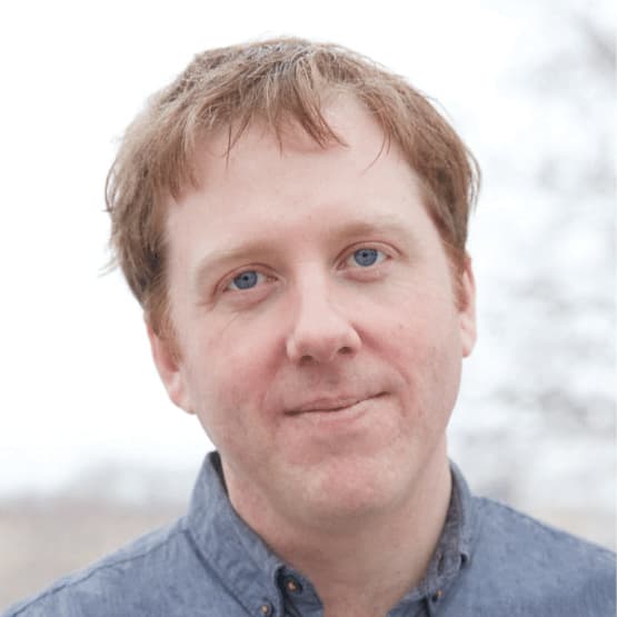 A man with light skin, short light brown hair, and blue eyes is looking at the camera with a slight smile. He is wearing a blue button-up shirt. The background is blurred and appears to be outdoors.