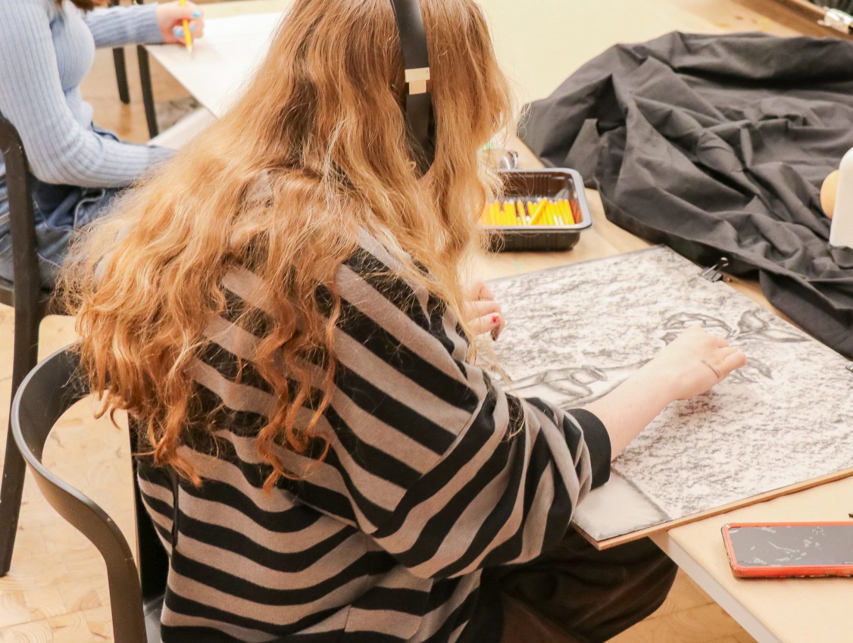 Several students sit around tables in an art classroom, sketching a still life arrangement of vases and fabric. One student in the foreground draws with charcoal, wearing a striped sweater and headphones.