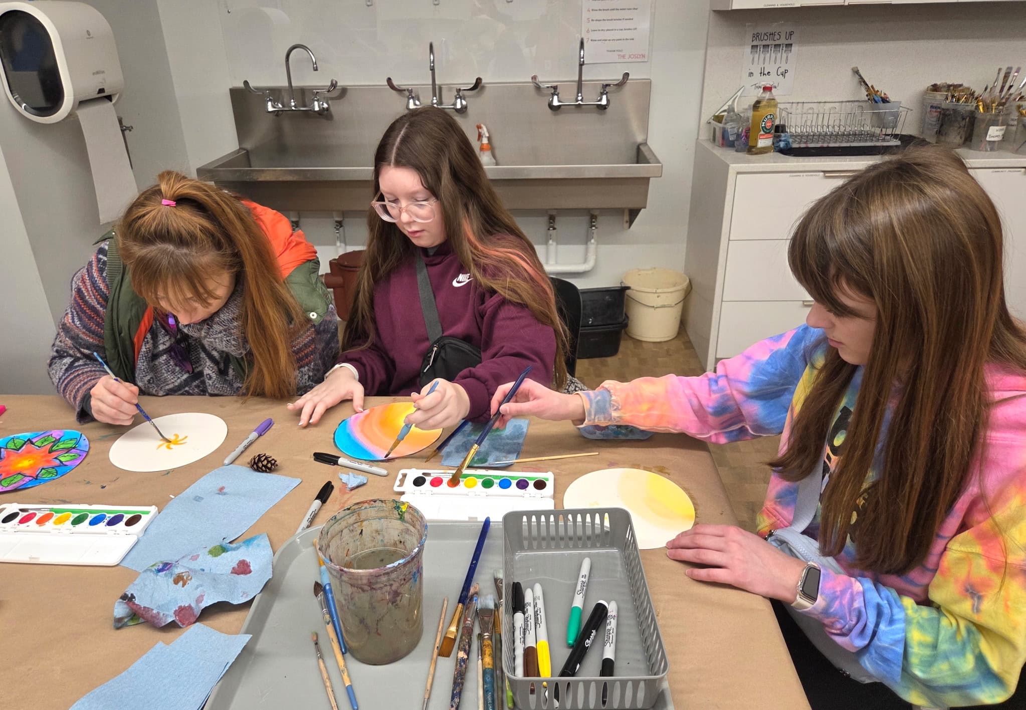 Three girls sit at a table painting round paper with watercolors in an art classroom. Art supplies, paint palettes, brushes, and paper towels are spread out on the table. A sink and shelves are visible in the background.
