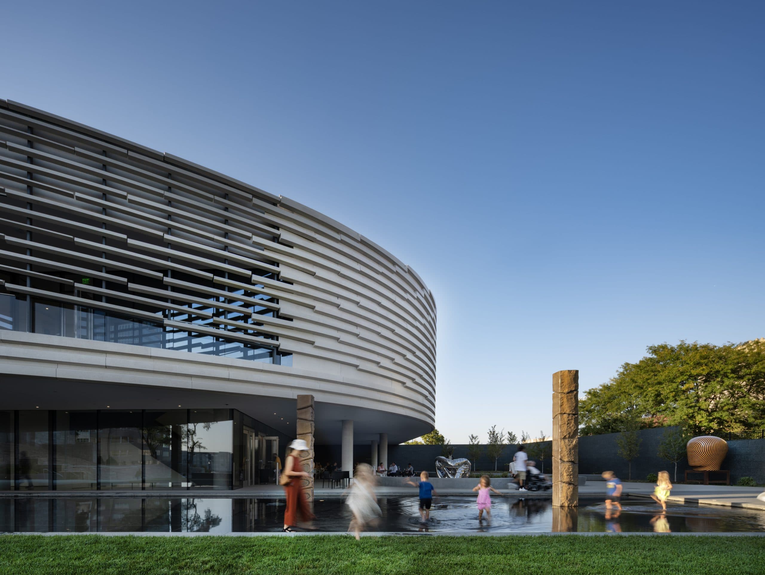 Children play in a shallow reflecting pool near a modern, curved building with horizontal slats on its facade, under a clear blue sky. Sculptural stone columns and greenery are visible in the background.
