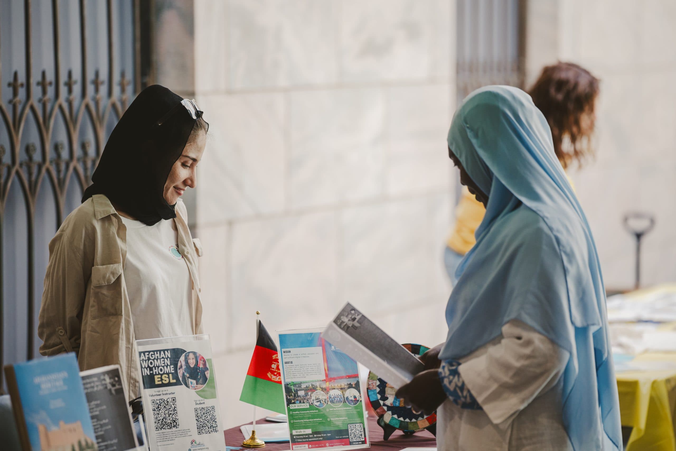 Two women wearing headscarves stand at a table covered with brochures, a small Afghan flag, and informational signs. One woman is reading a flyer while the other smiles. Other tables are blurred in the background.