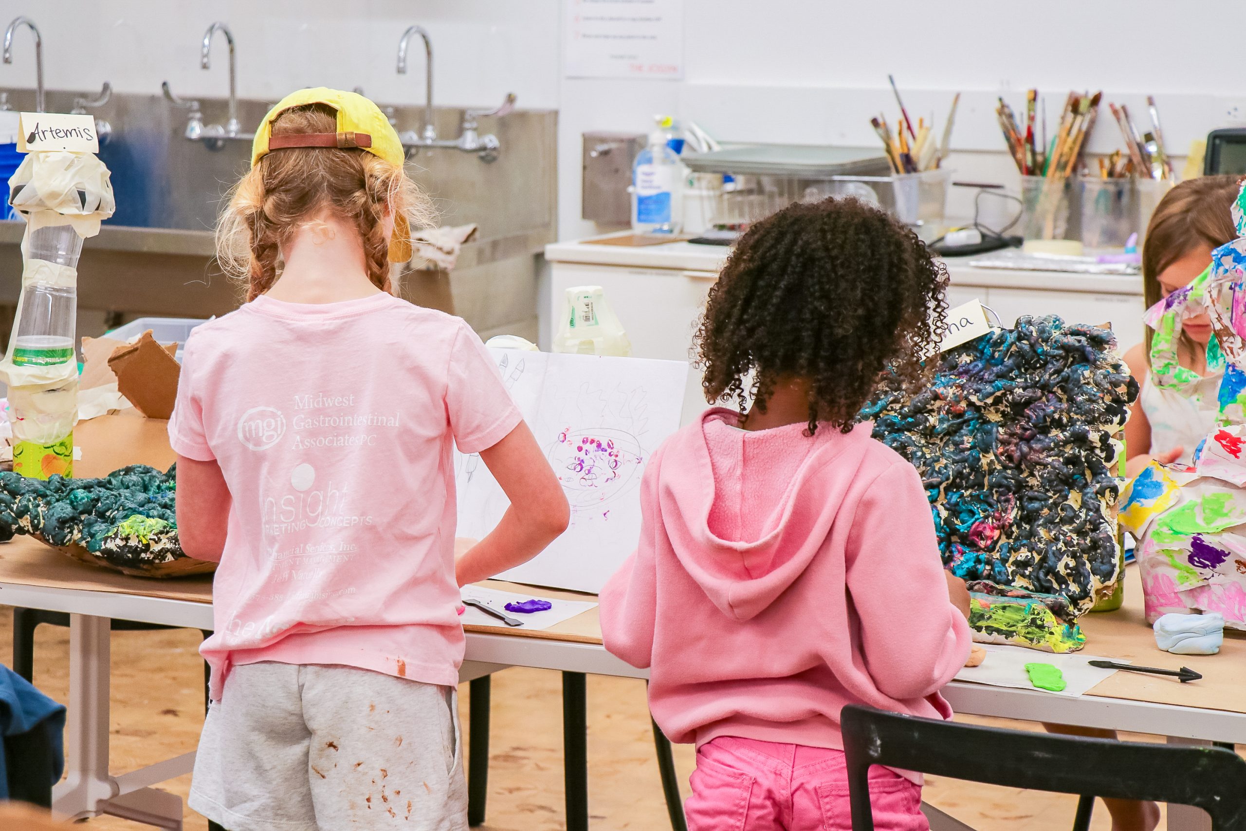 Two young children stand at a table in an art classroom, engaged in a creative project with colorful materials and textured sculptures. Art supplies and tools are scattered on the table and countertops in the background.
