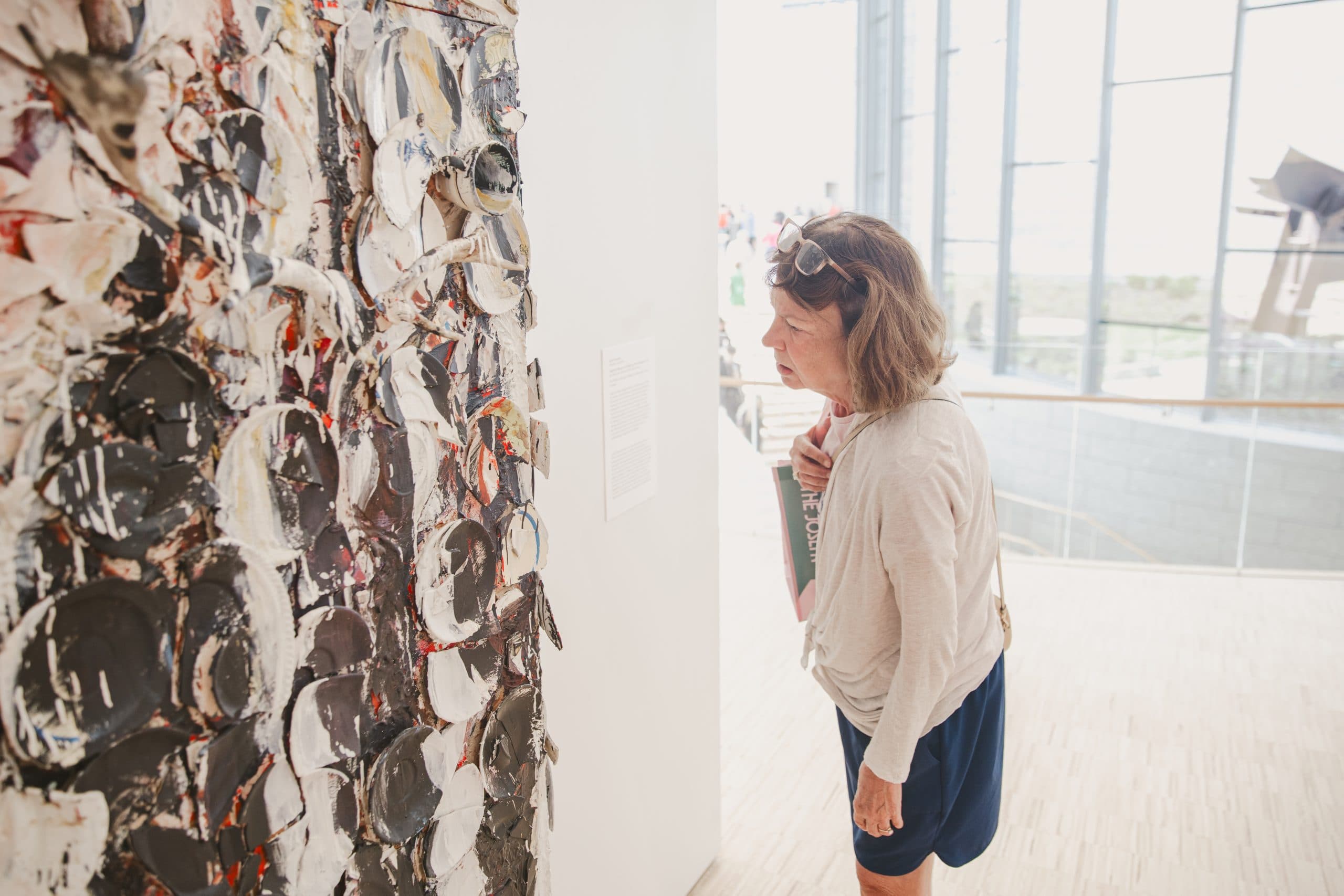 A woman closely examines a textured, abstract painting with circular patterns in a bright, modern art gallery filled with natural light.