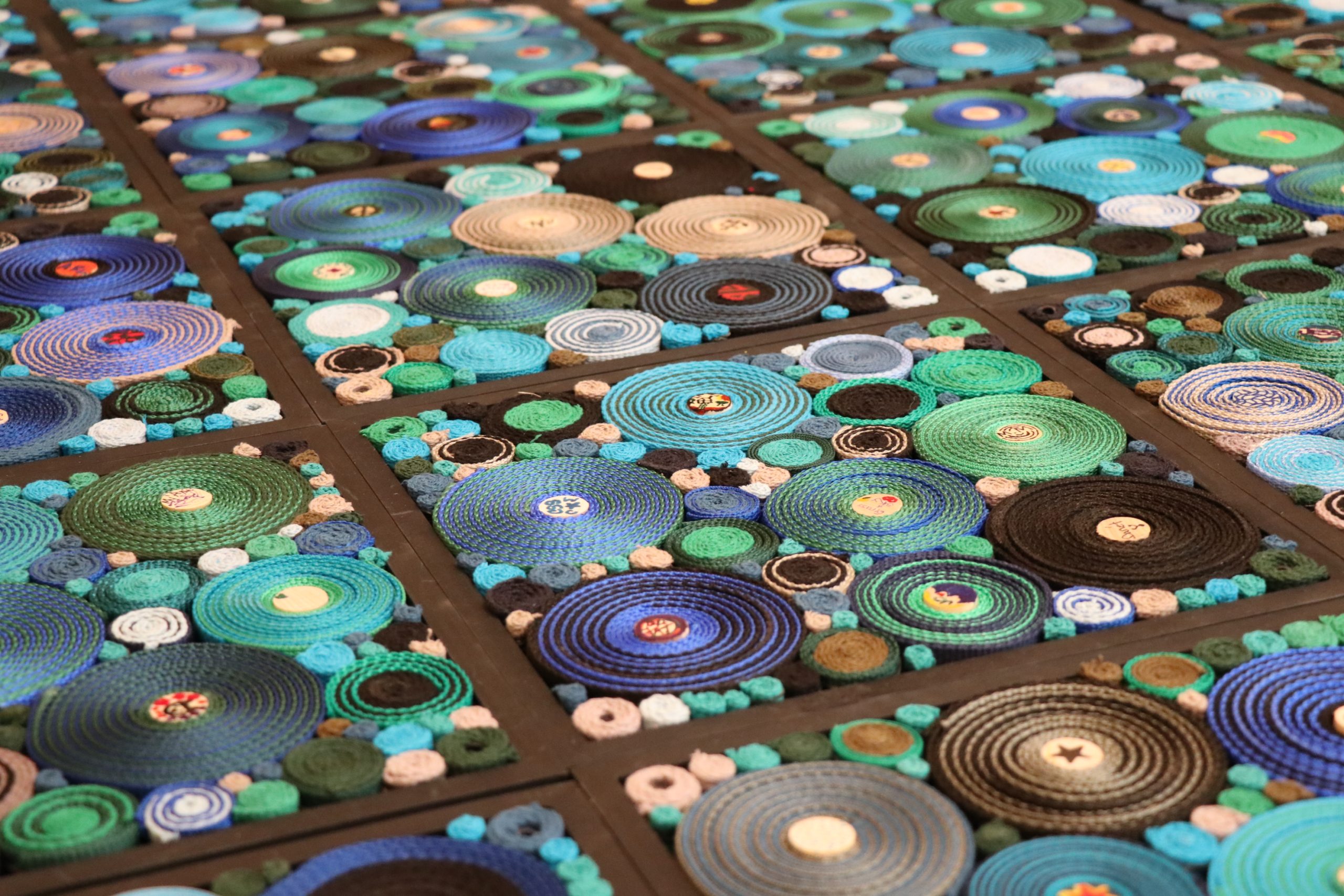 A close-up of a colorful mosaic art piece featuring concentric circles in various shades of blue, teal, and green, arranged in a grid pattern with textured, layered surfaces.