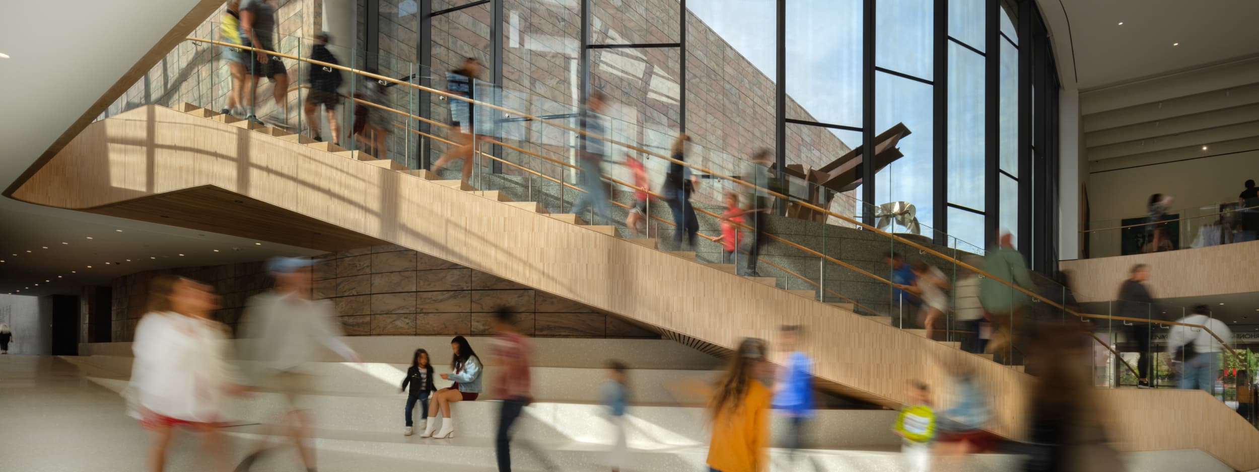 People walk up and down a wide wooden staircase inside a modern building with large windows and stone walls; others move or sit in the bright, open lobby area below.