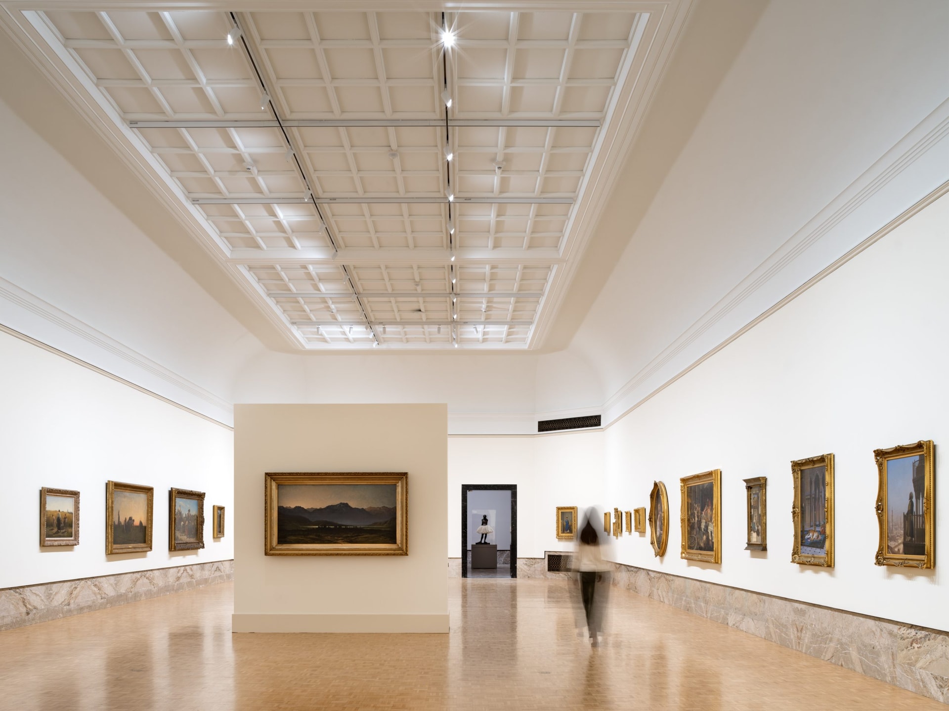 A spacious art gallery with a high, coffered ceiling and walls lined with framed paintings. A blurred figure walks on the polished wooden floor, and a statue is visible through a doorway at the far end.