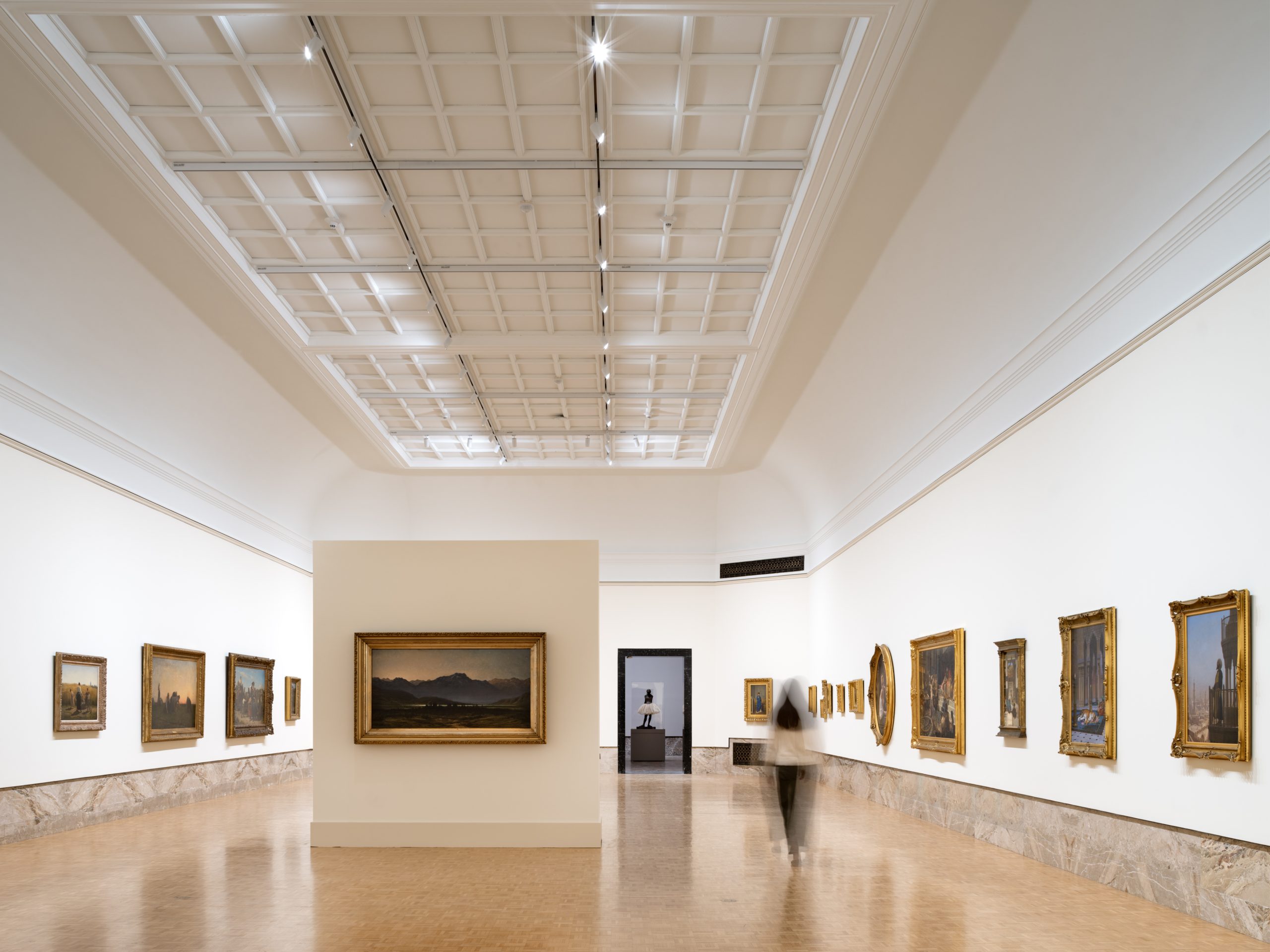 A spacious art gallery with a high, coffered ceiling and walls lined with framed paintings. A blurred figure walks on the polished wooden floor, and a statue is visible through a doorway at the far end.