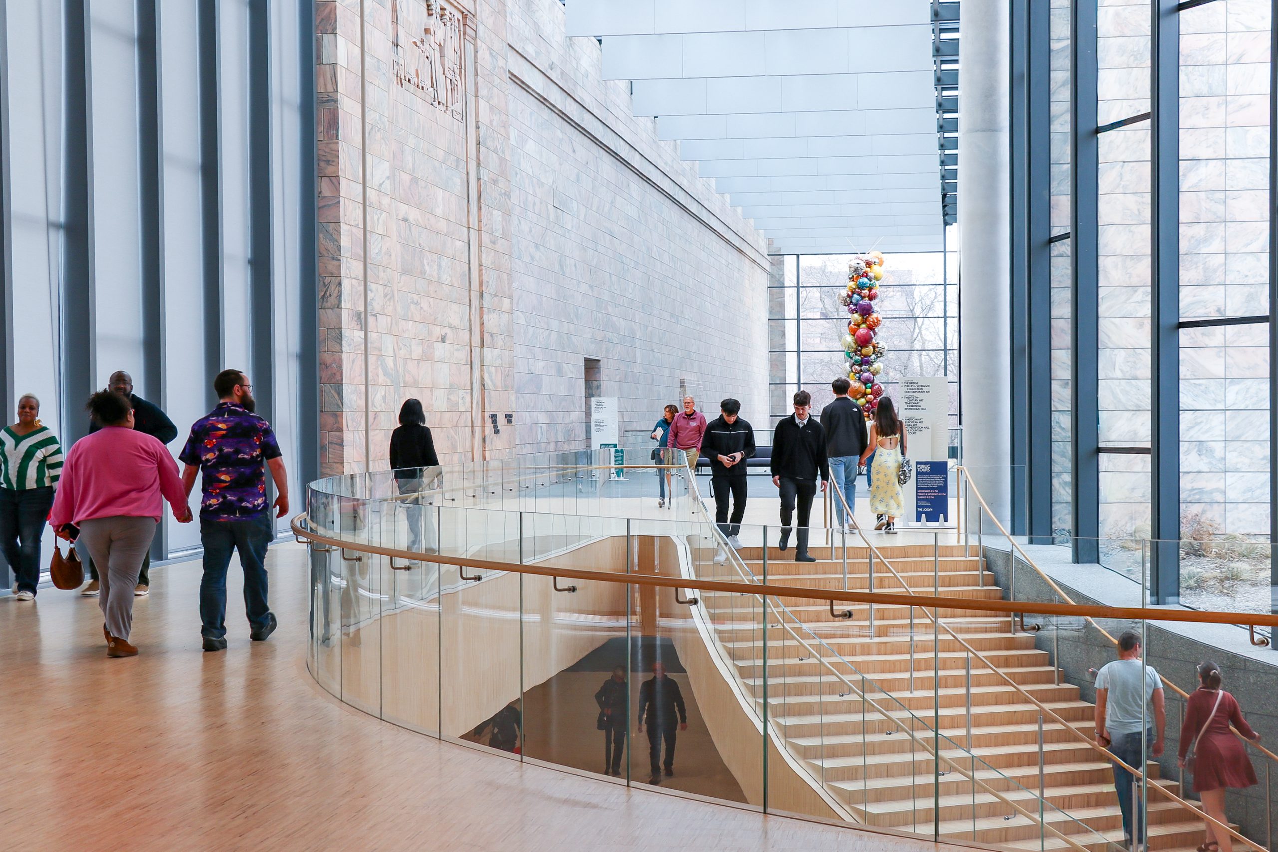People walk through a spacious, modern museum hallway with tall glass windows, a stone wall, and a colorful hanging sculpture in the background. A staircase curves downward in the center.