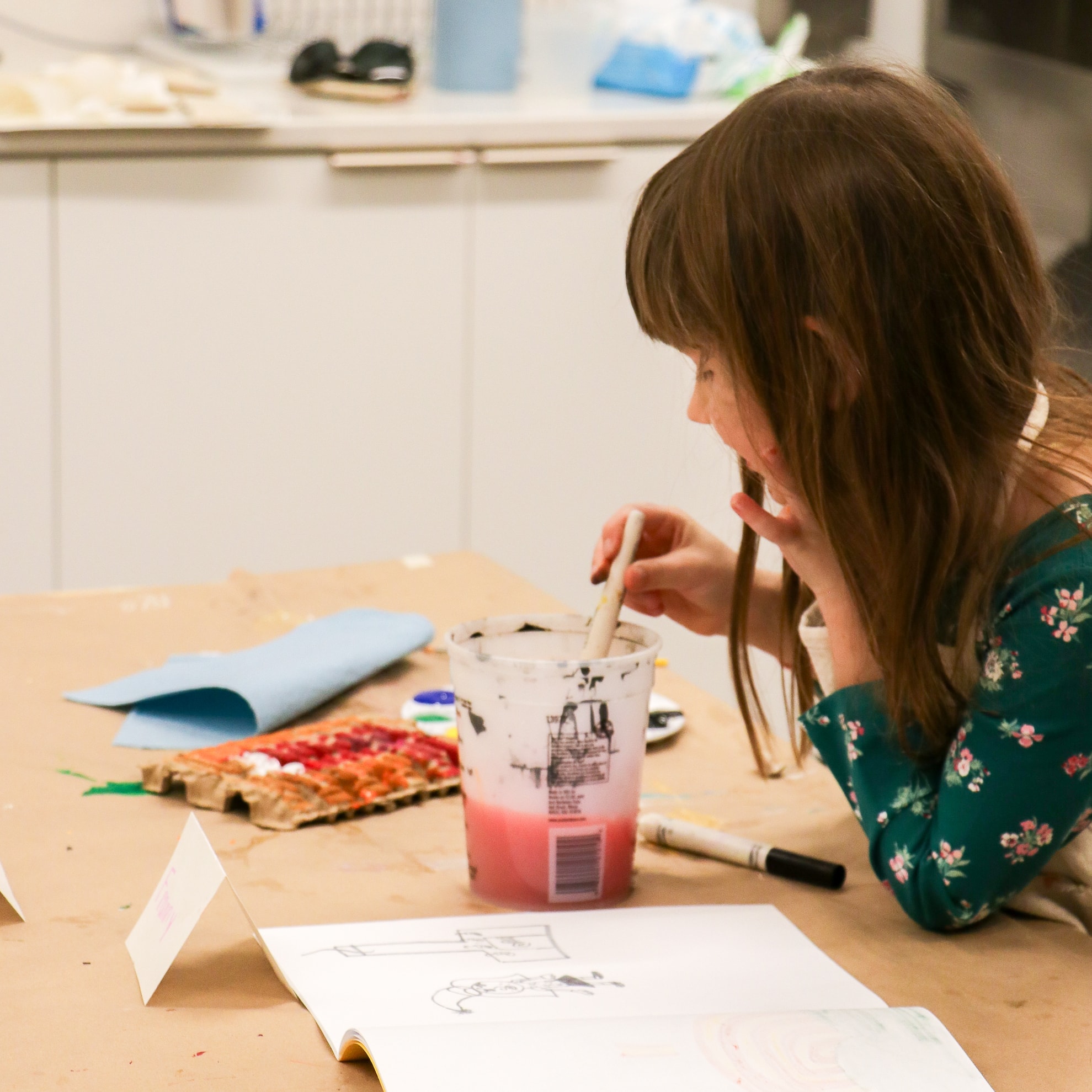 A young girl in a green floral dress dips a paintbrush into a cup of water while sitting at a table covered with art supplies, paint palettes, and paper in a bright room.