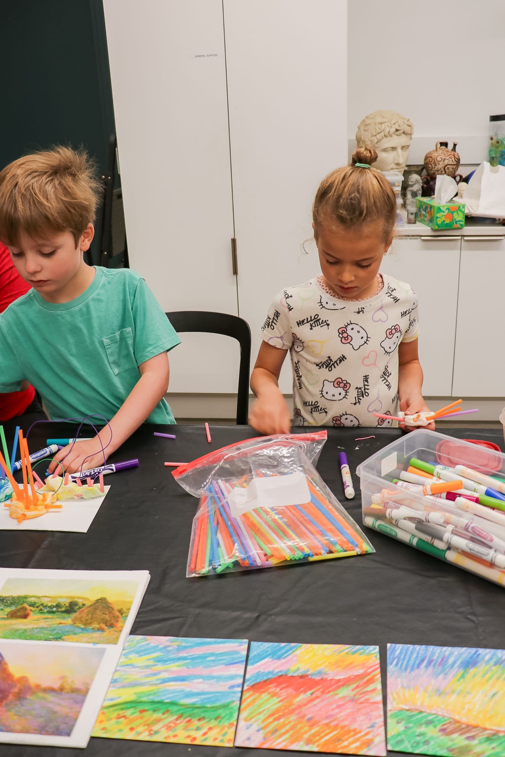 Two children sit at a table covered with colorful markers, pencils, and artwork. They are focused on drawing and creating crafts, with art supplies scattered around them. Brightly colored finished drawings are displayed on the table.