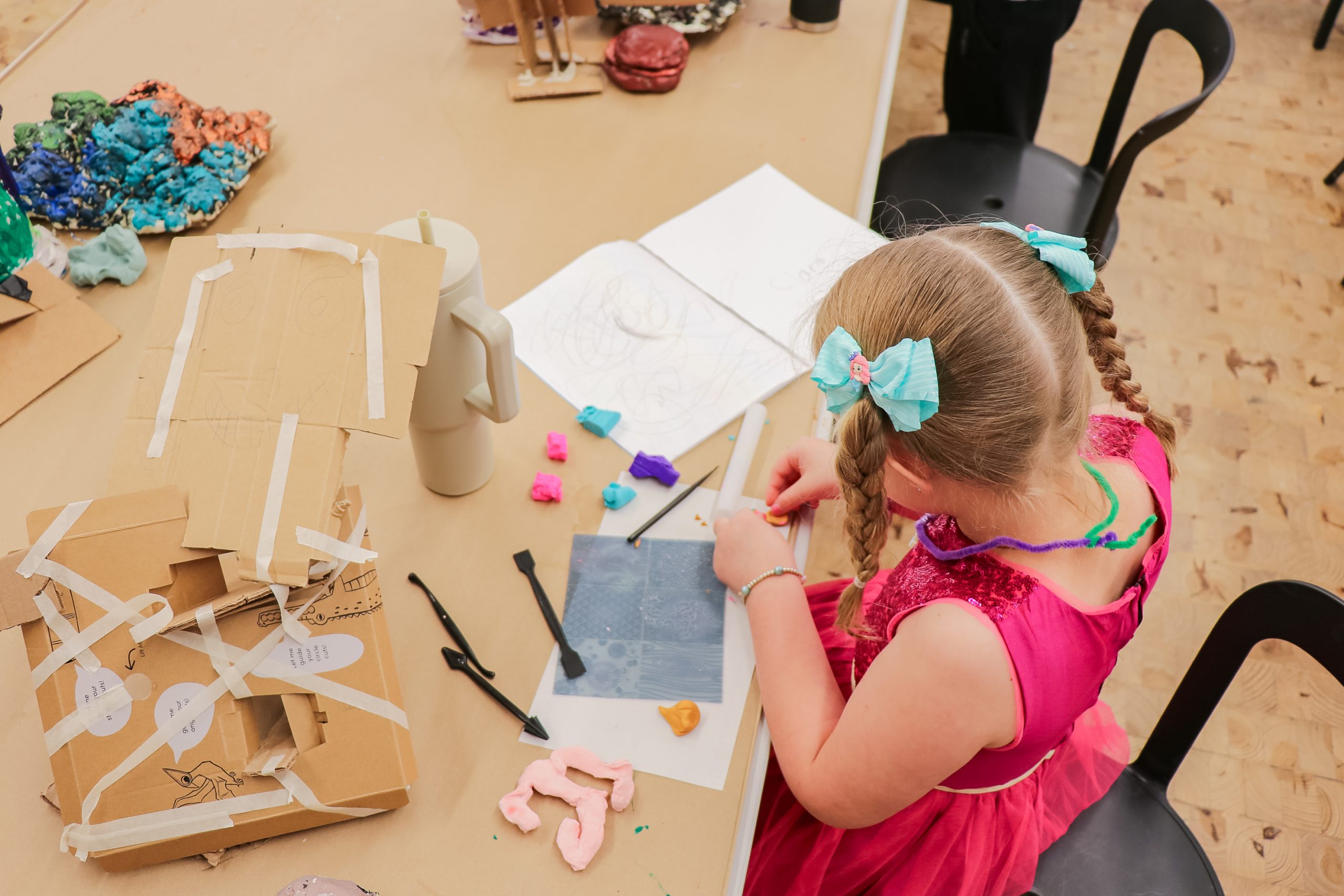A young girl with braided hair and blue bows, wearing a pink dress, sits at a table working on an art project with paper, clay, and cardboard. Art supplies and colorful clay pieces are scattered on the table.