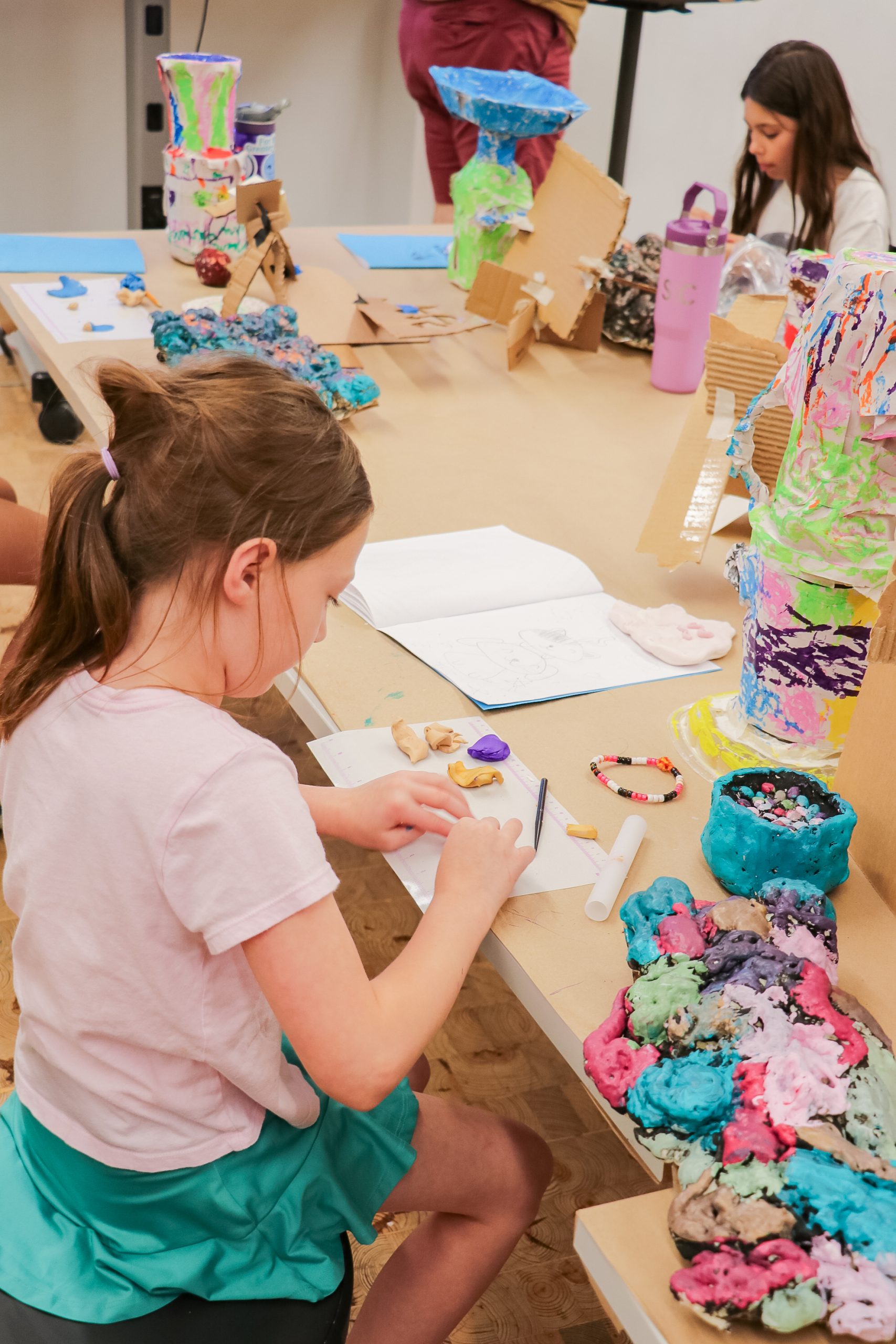 A young girl sits at a table drawing in a sketchbook surrounded by colorful clay sculptures and art supplies, while another child works in the background.