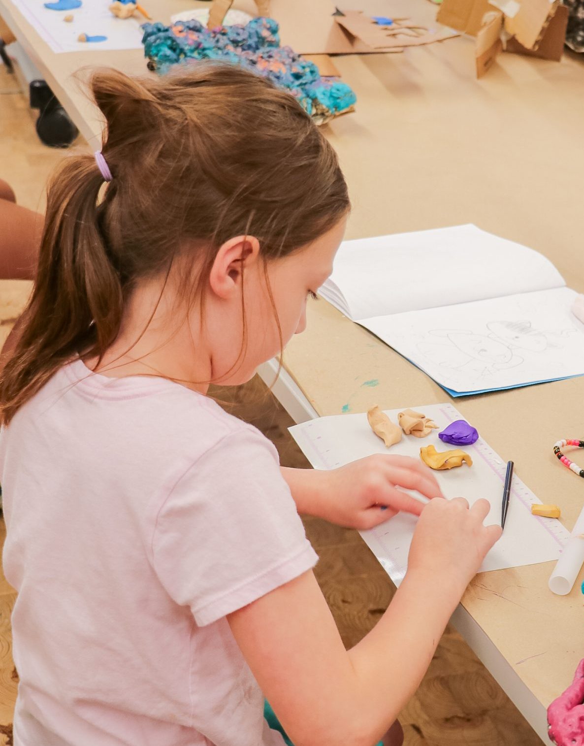 Young girl sits at a table drawing in a sketchbook, surrounded by colorful clay art projects and sculptures. Another child works in the background, also focused on creative activities.