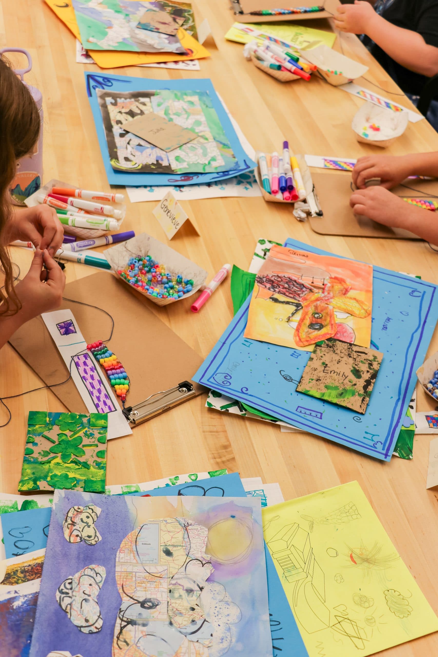 Children’s hands work on colorful art projects at a table covered with drawings, paintings, markers, beads, and craft supplies during a creative arts and crafts activity.