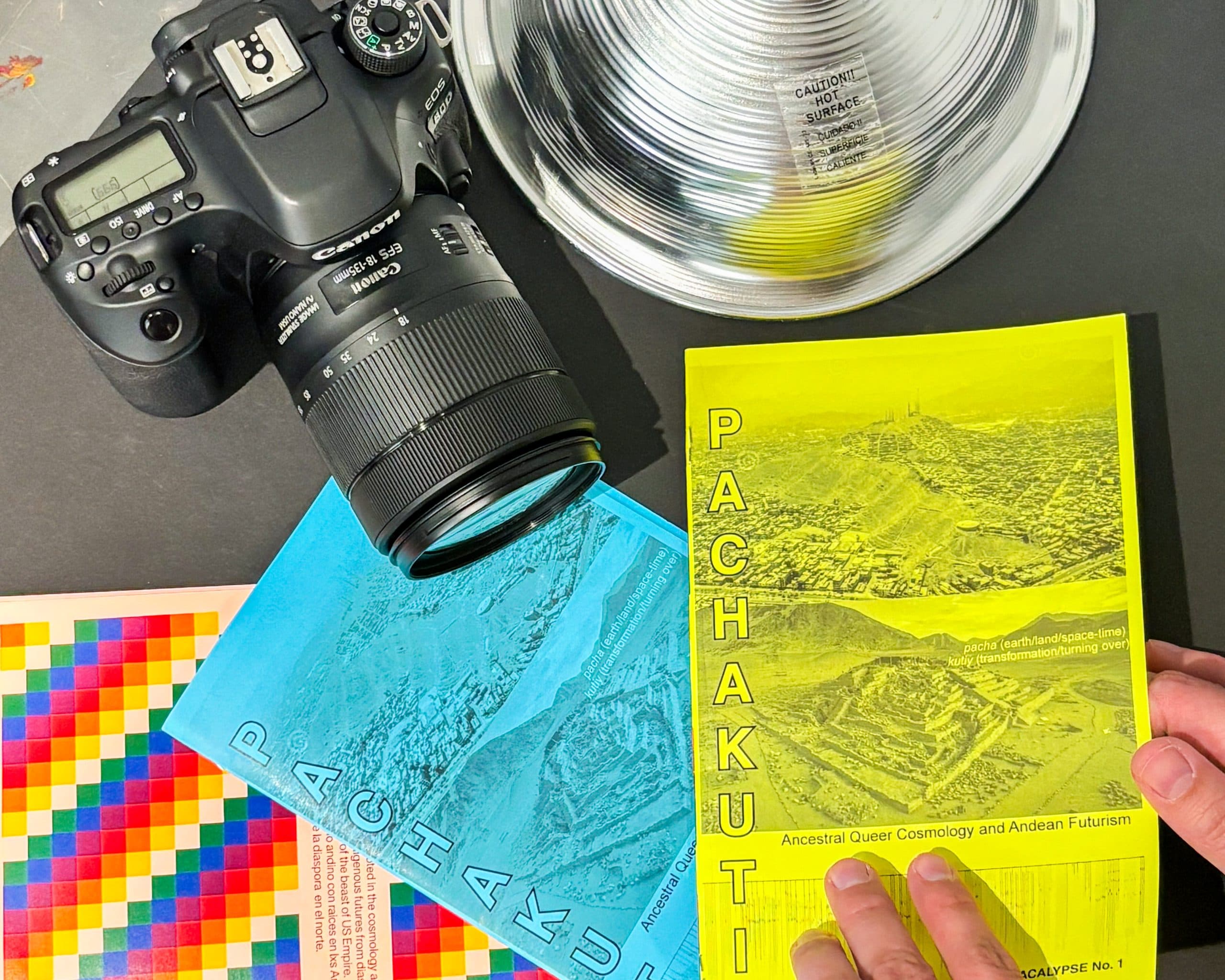 A hand holds a yellow book titled "PACHAKUTI" near a blue version of the same book, a DSLR camera, a glass bowl, and a colorful pixelated sheet on a black surface.