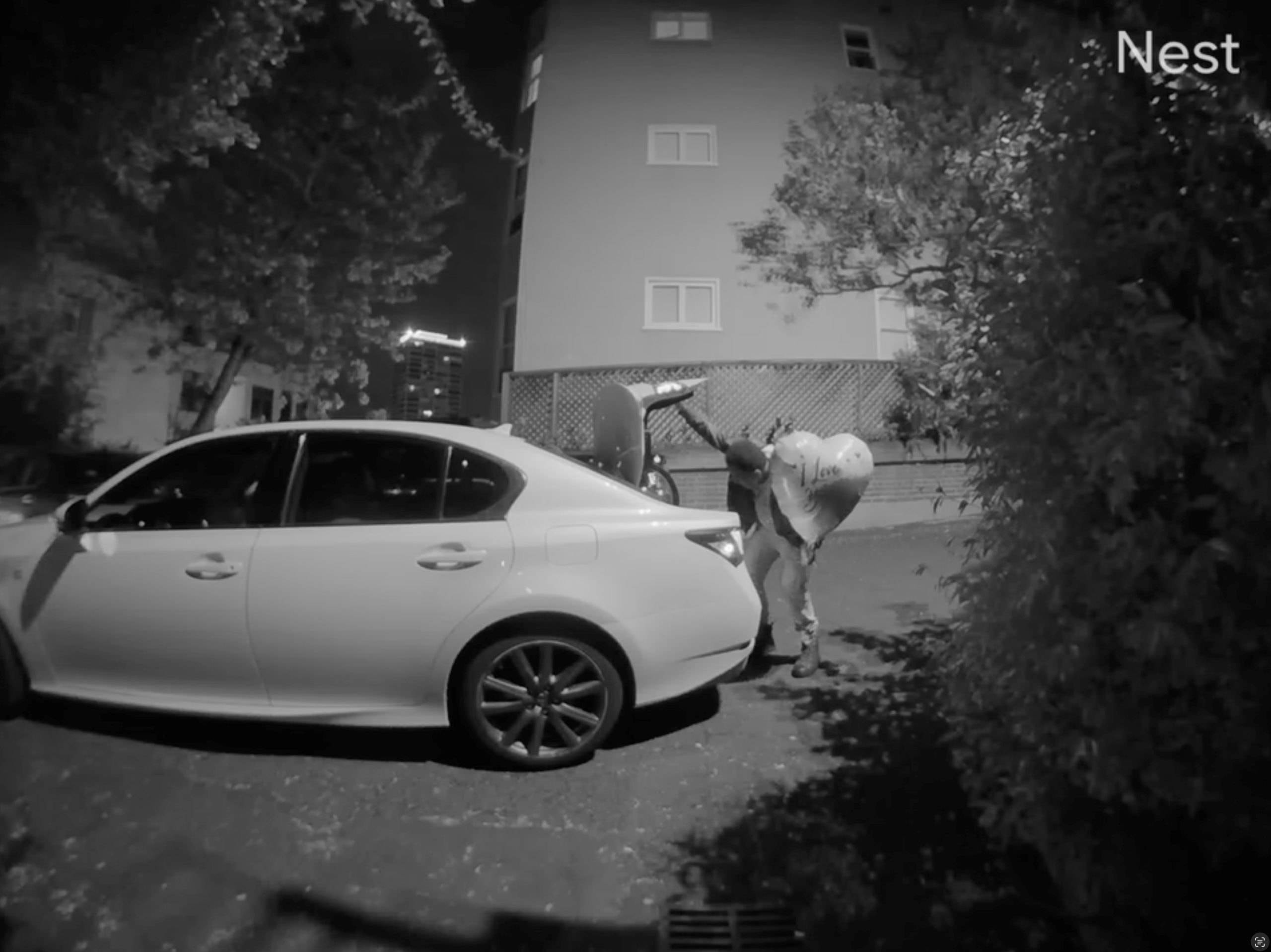 A person at night is bending over the trunk of a white car in a parking area, partially hidden by bushes. The scene is captured in black and white by a Nest security camera.