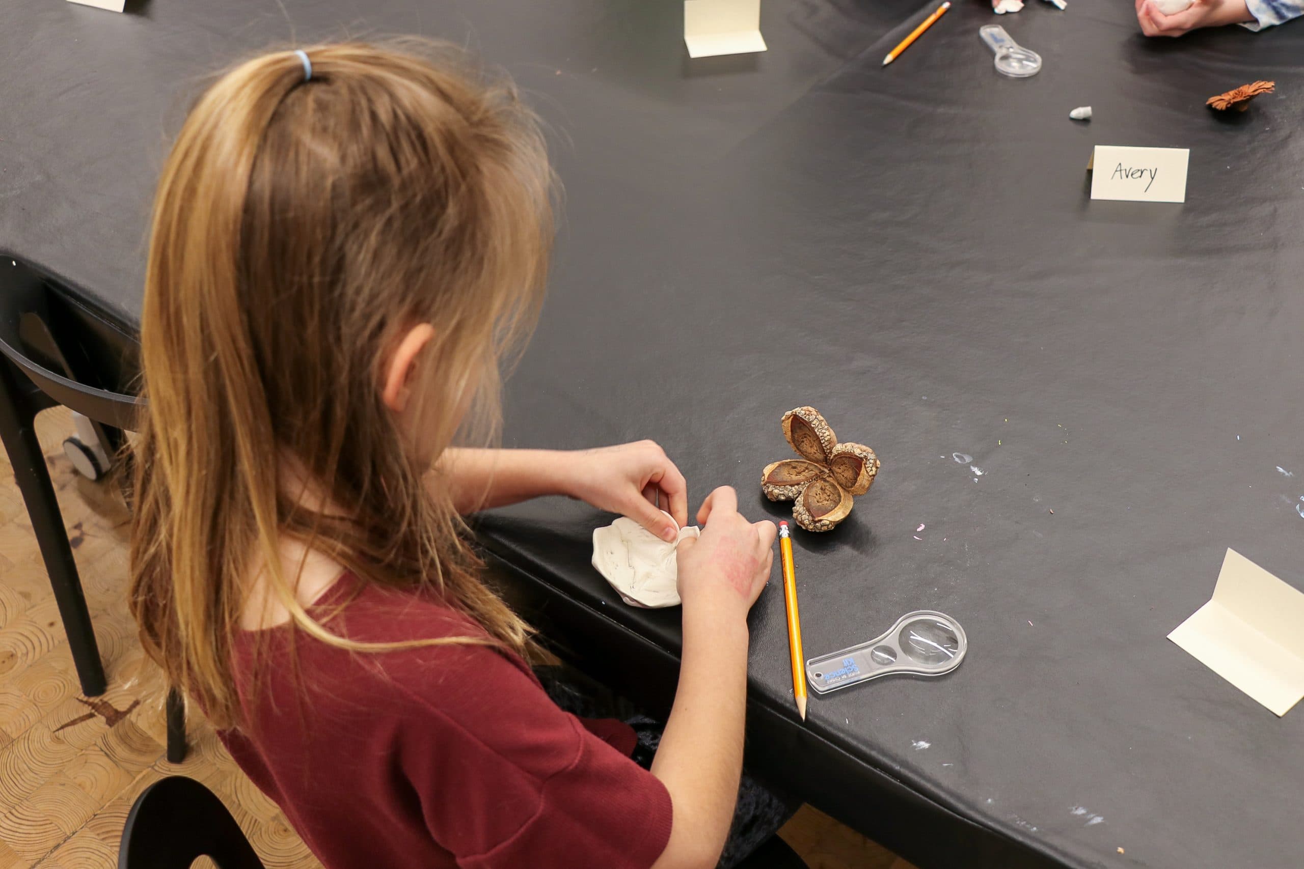 A child with light brown hair sits at a table, working with clay and natural materials. A pencil, magnifying glass, and name cards are on the black tablecloth in front of her.