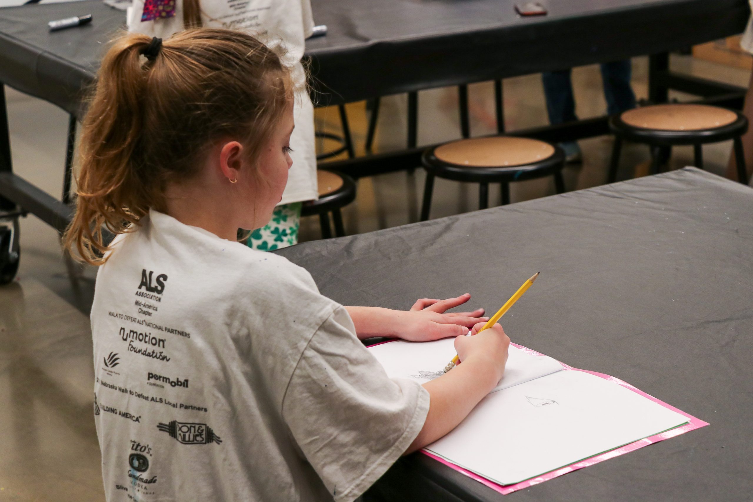 A young girl sits at a black-covered table, drawing in a sketchbook with a pencil. She is wearing a white t-shirt with logos on the back, and there are stools and another child in the background.