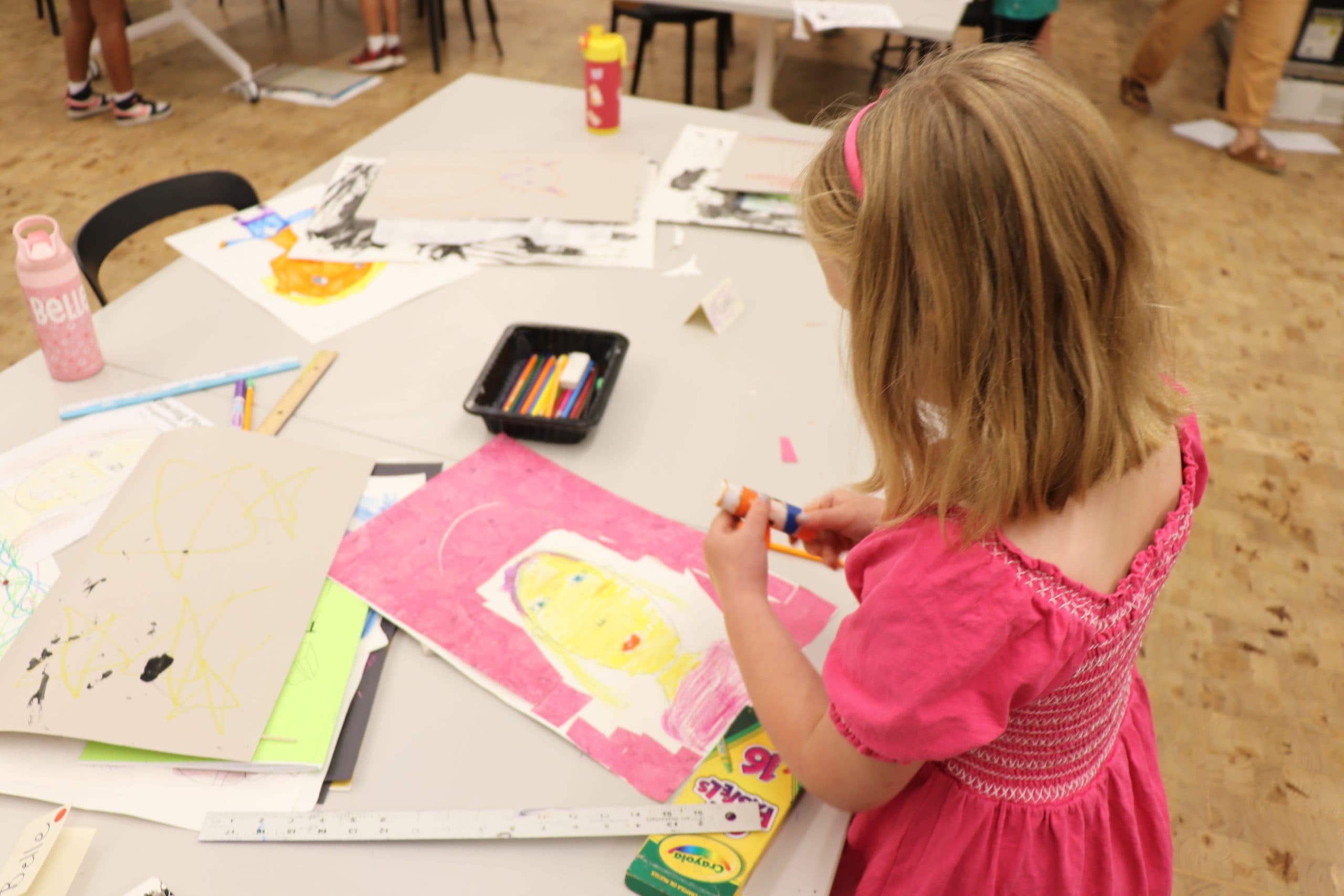 A young girl in a pink dress uses a glue stick to work on a colorful drawing at a table covered with art supplies, drawings, and crayons in a classroom or art studio setting.