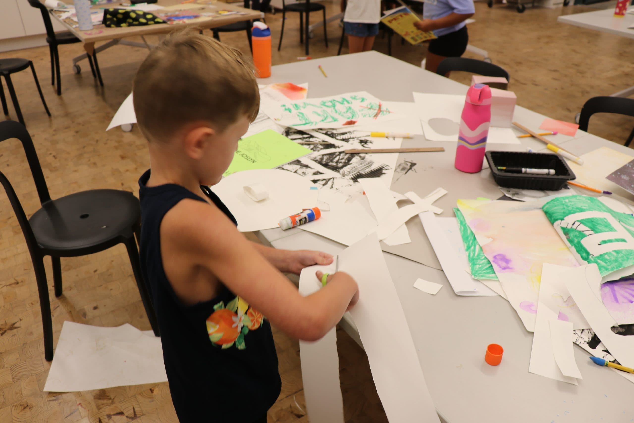 A young boy stands at a messy table covered with art supplies, paper, markers, and glue, focused on cutting a piece of white paper during a craft activity.