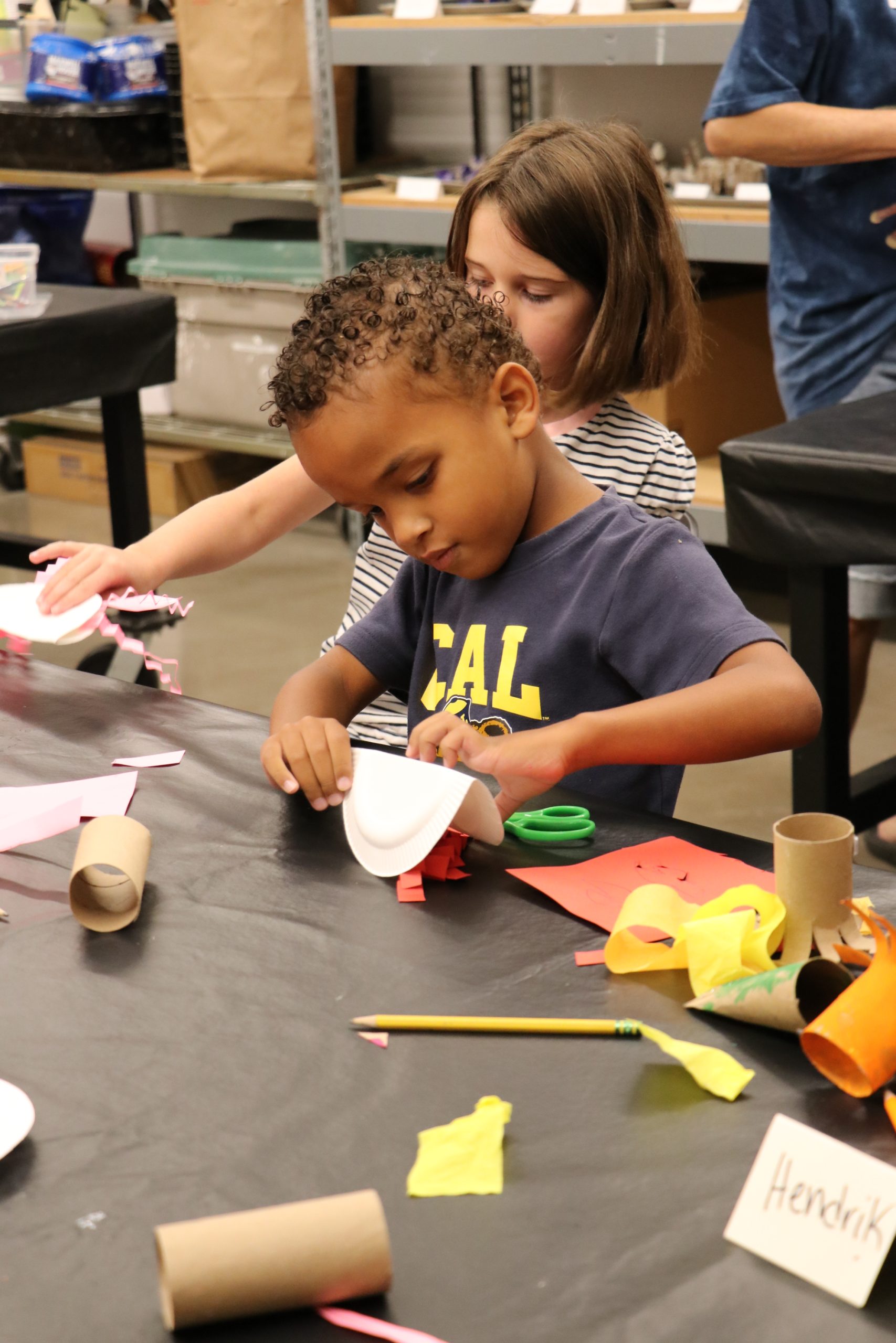 Two children sit at a table doing crafts with paper, scissors, and other materials. The boy in front focuses on gluing a white paper plate, while the girl behind him works on her own project. Craft supplies cover the table.