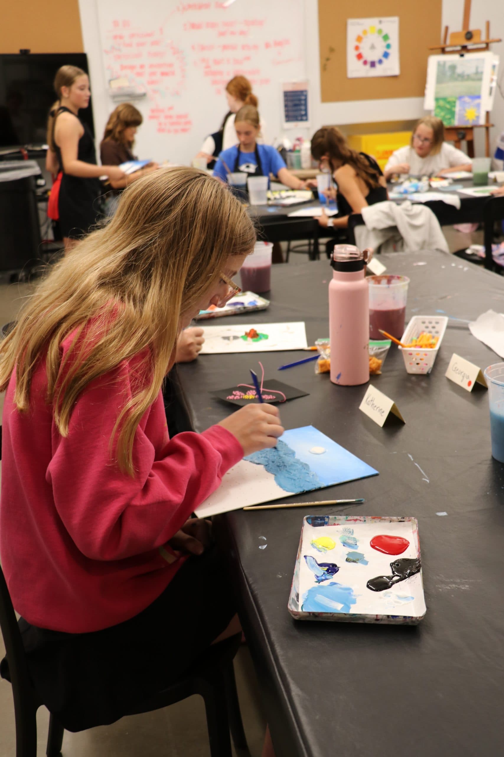 A group of children are sitting at tables in an art classroom, painting on canvases. A girl in the foreground is focused on her artwork, with paint supplies and a water bottle nearby. Other kids are working in the background.