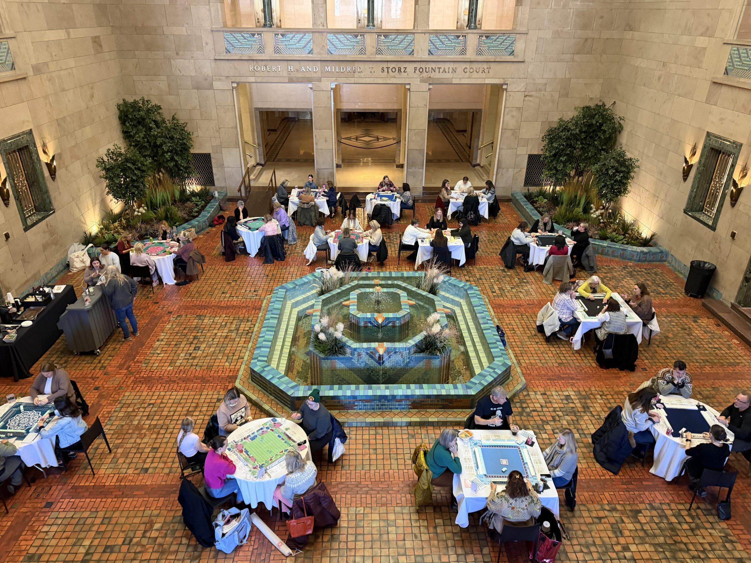 People sit in groups at round tables playing board games in a large, elegant hall with a decorative fountain in the center, surrounded by plants and tiled floors. The atmosphere appears lively and social.
