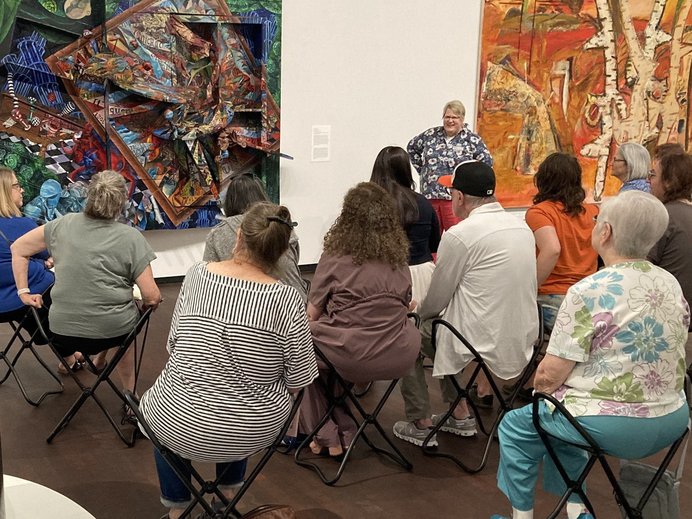 A group of people sit on folding stools, listening to a woman speak in front of two colorful abstract paintings in an art gallery.