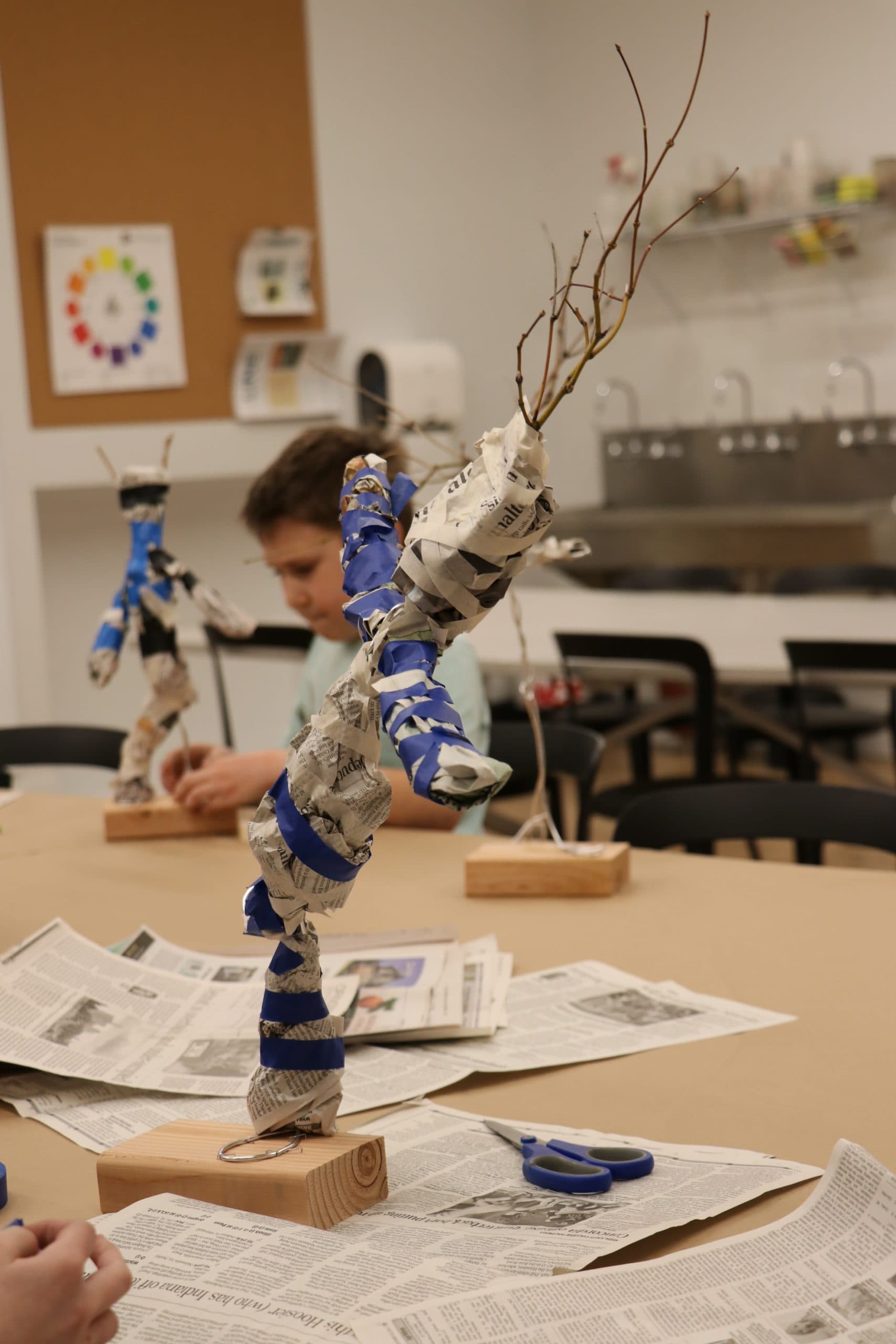 A child works at a table creating a sculpture from twisted newspaper, blue tape, and branches, with another similar sculpture in the background. Art supplies and newspaper are scattered on the table.