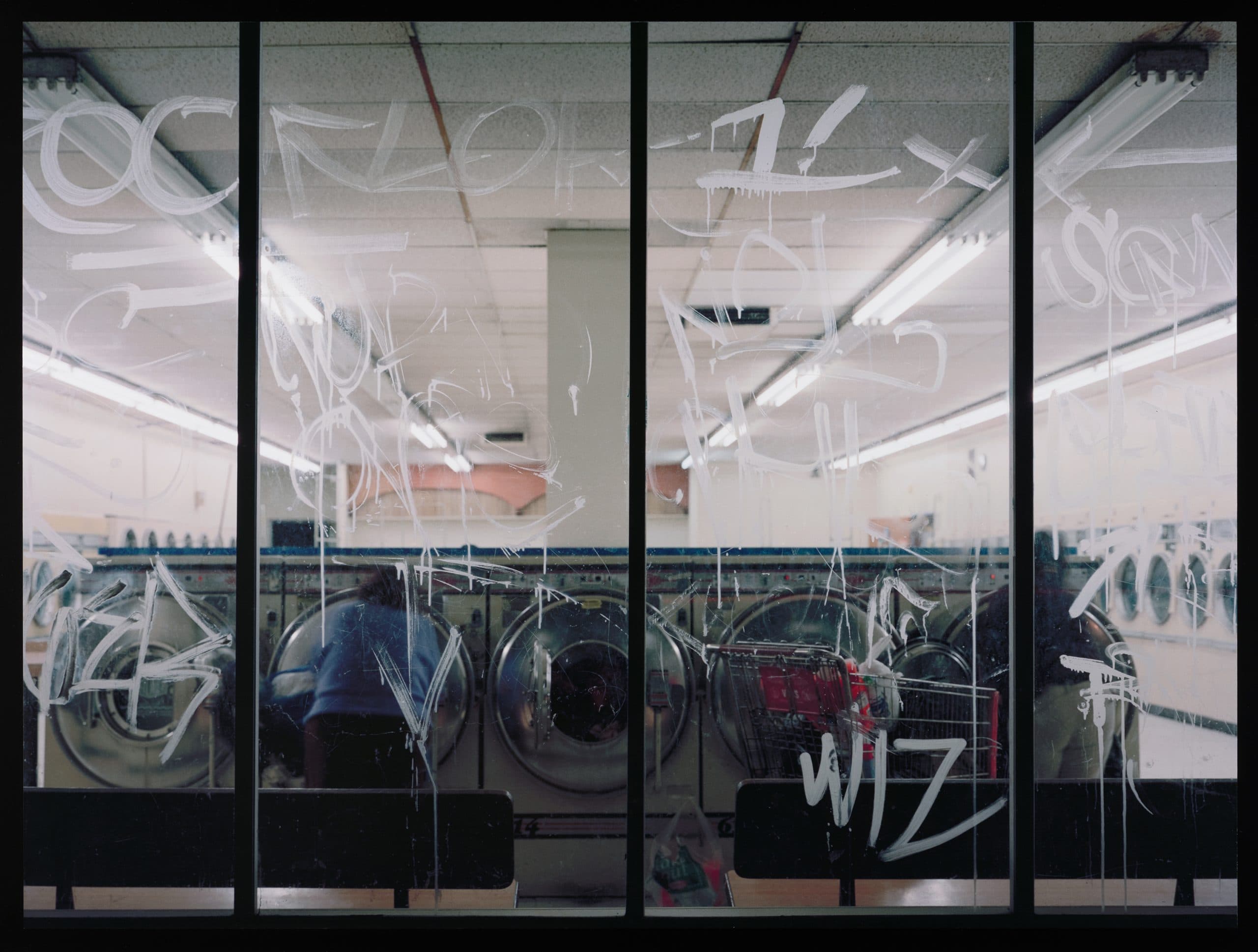 A laundromat viewed through large glass windows covered in white graffiti, with washing machines and a person faintly visible inside under fluorescent lighting.