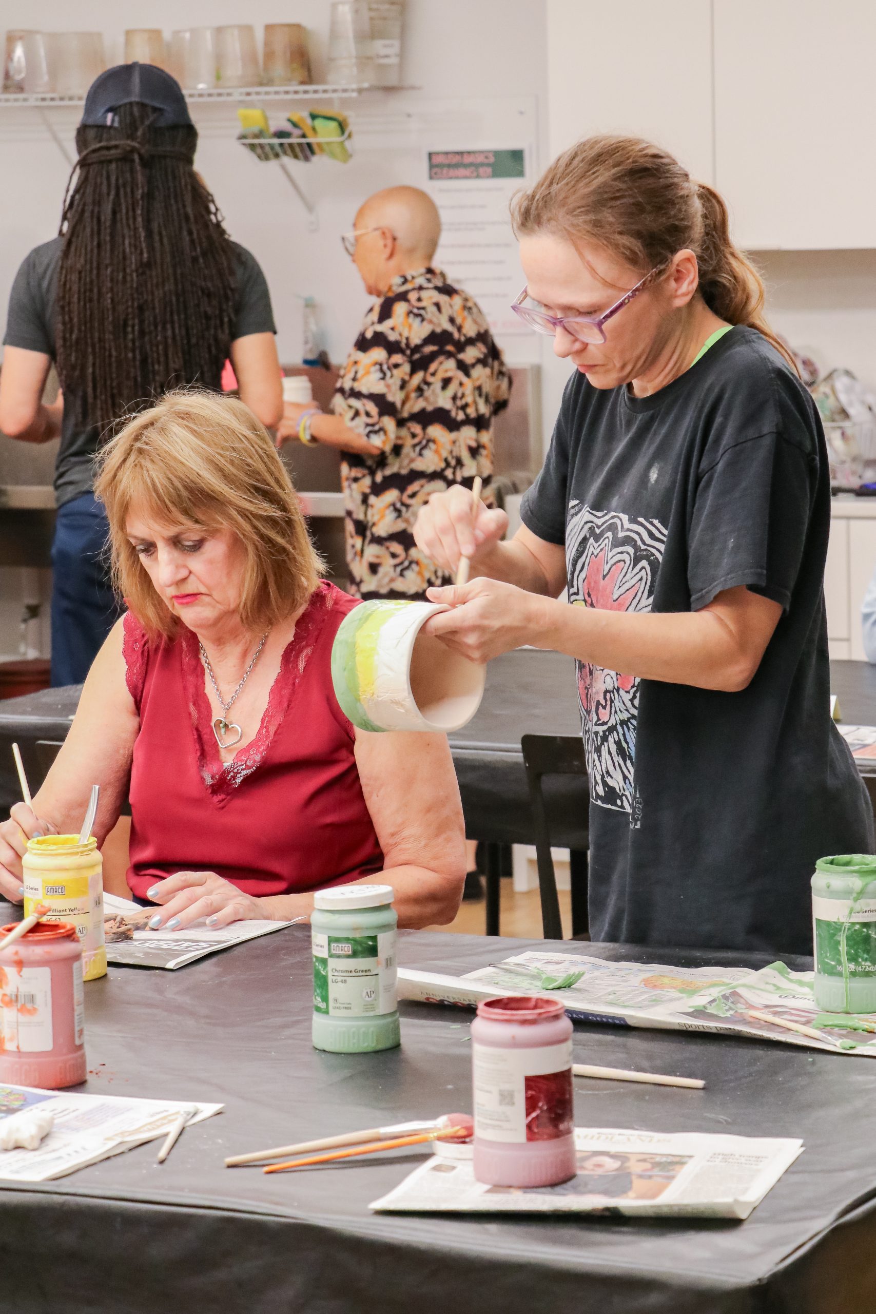 Several people are in a bright art studio; one woman paints pottery, while another, seated, paints a ceramic piece. Jars of paint and brushes are scattered on the table. Other people work in the background.