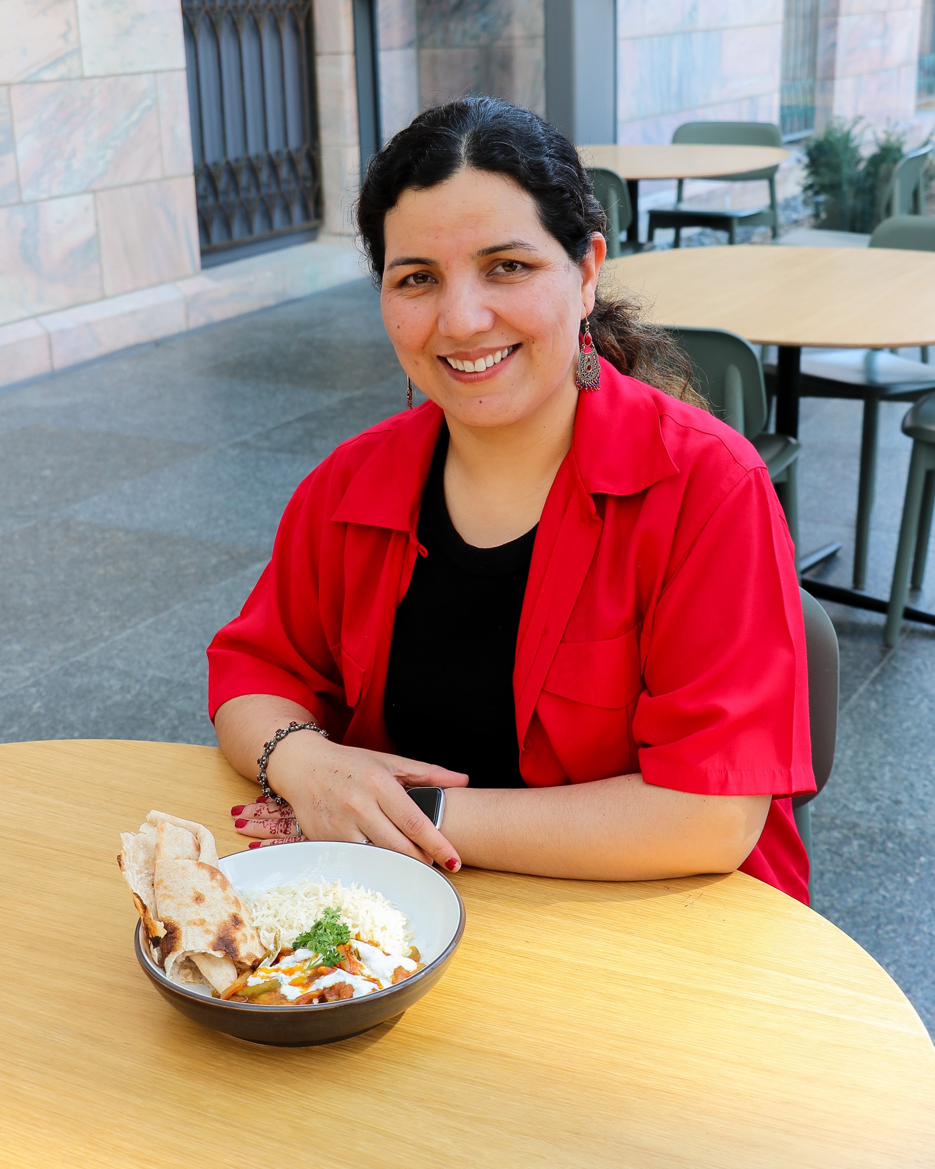 A woman with curly dark hair, wearing a red shirt and black top, sits at a table outdoors, smiling. In front of her is a bowl of food with rice, sauce, and flatbread. Empty tables and chairs are seen in the background.