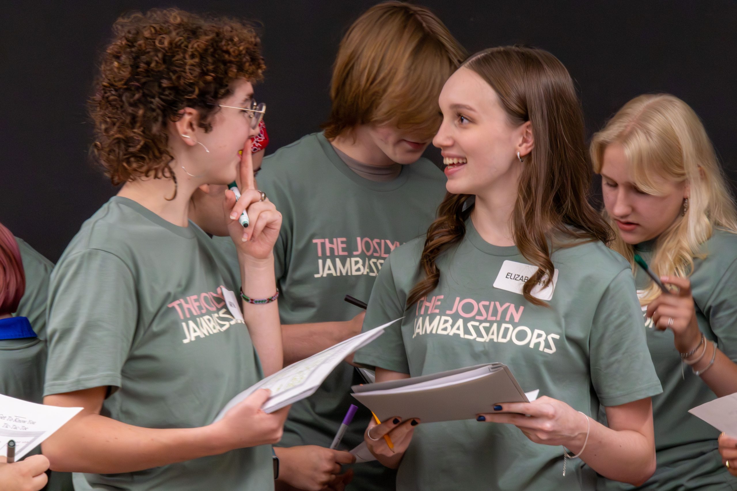 A group of teens wearing matching "The Joslyn Jambassadors" t-shirts stand together, holding papers and pens, engaged in lively conversation and smiling. One girl, with a name tag reading "Elizabeth," smiles at a peer.